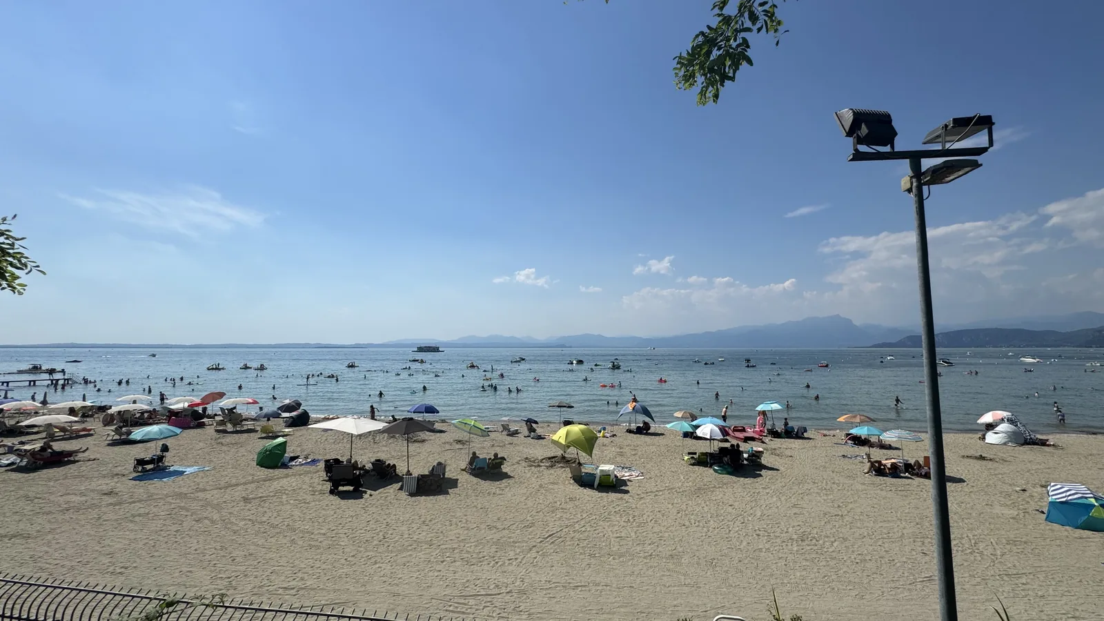 Breiter heller Sandstrand am Camping La Quercia in Lazise mit flachem Wasser voller Badender und der Bergkette am Gardasee im Hintergrund