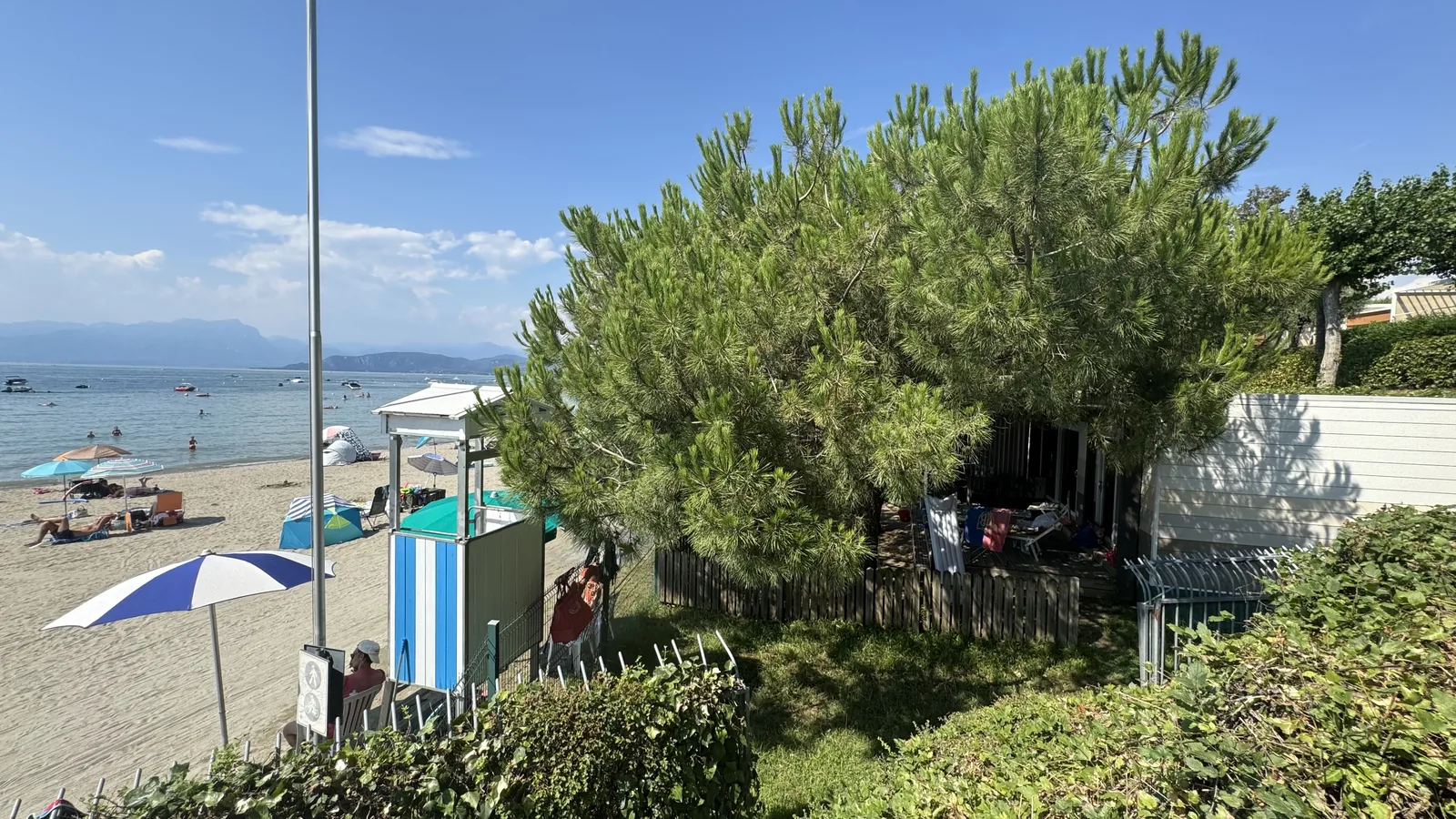 Rettungsschwimmer-Kanzel und Verleih-Häuschen unter einer Schirmpinie am Strandzugang, dahinter ein Mobilheim