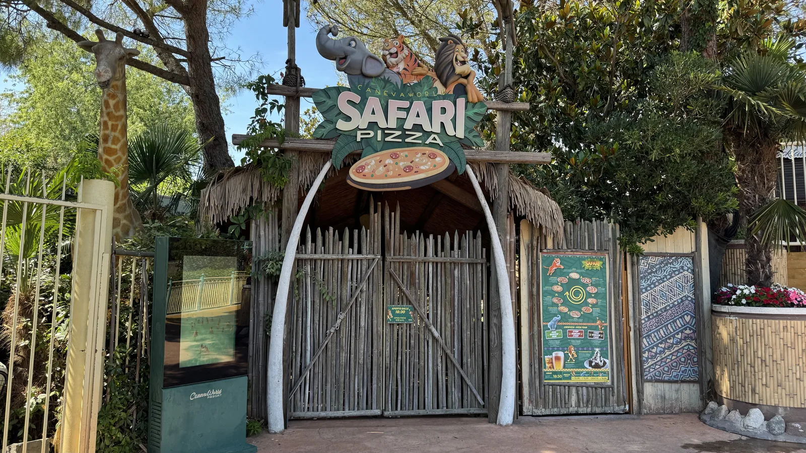 Geschlossenes Bambustor mit dem Schriftzug Safari Pizza, darüber gemalte Tierköpfe und eine Pizza-Attrappe, links eine Giraffenfigur
