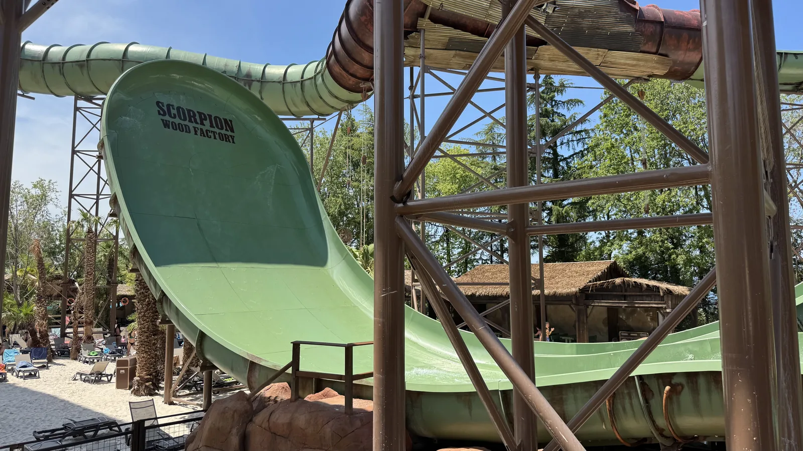Grüne Rutschenschale mit dem Schriftzug Scorpion Wood Factory, eingefasst von einem offenen braunen Stahlgerüst, dahinter Palmen und Liegen