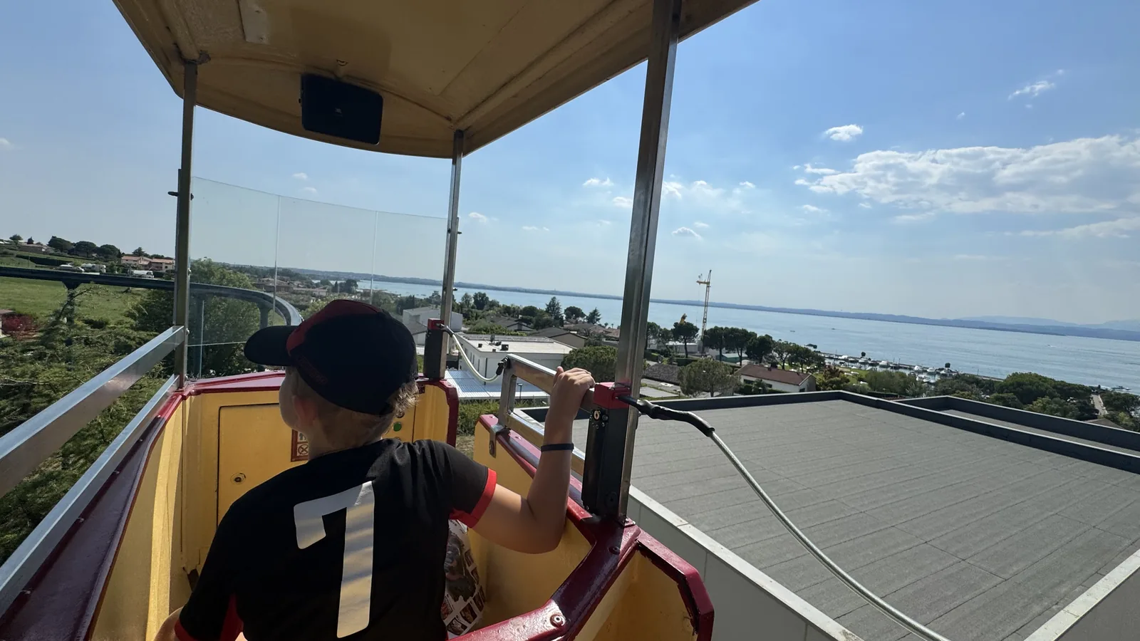 Kind von hinten im gelb-roten Wagen einer erhöht fahrenden Bahn, im Hintergrund der Gardasee mit Uferorten und Bergen