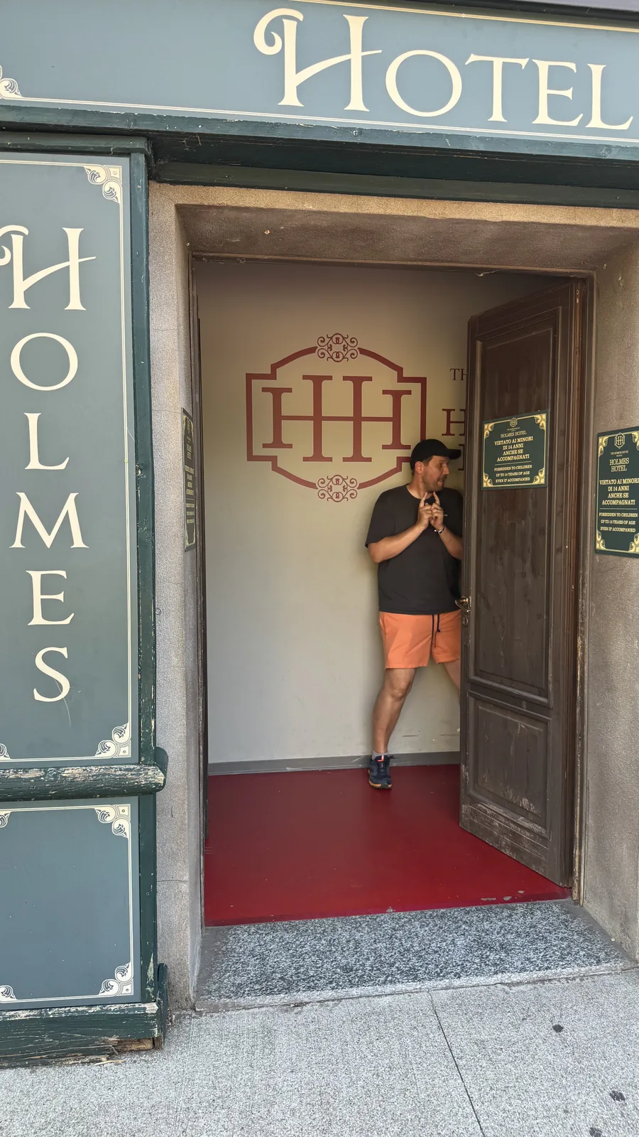Eingang zum Holmes Hotel im Movieland Park: dunkelgrüne Fassade, offene Holztür, roter Teppich im Flur, daneben ein grünes Hinweisschild mit der Altersgrenze