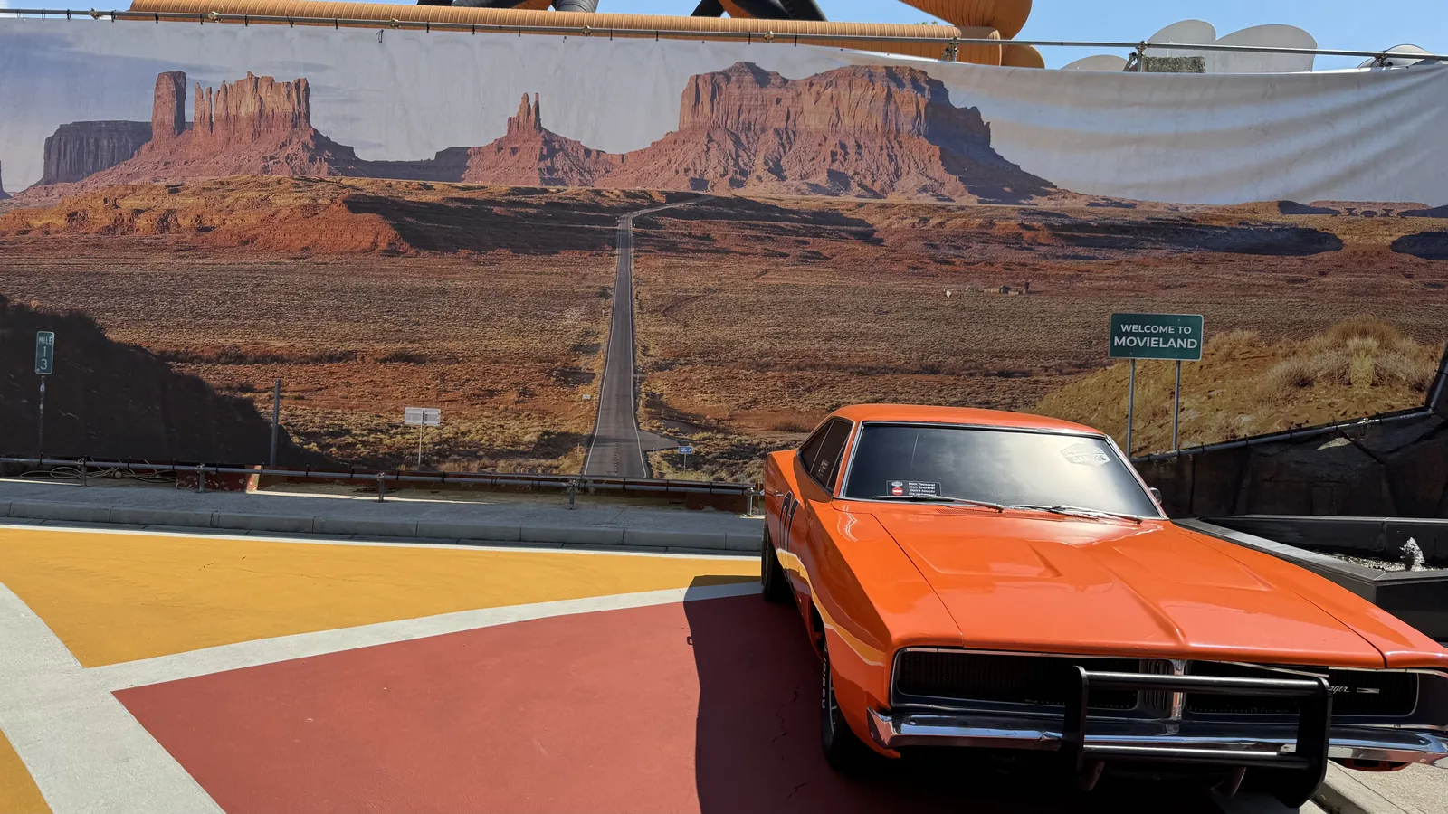Orangefarbener amerikanischer Straßenkreuzer vor einer großformatigen Fotokulisse des Monument Valley im Movieland Park, daneben ein Straßenschild mit der Aufschrift Welcome to Movieland