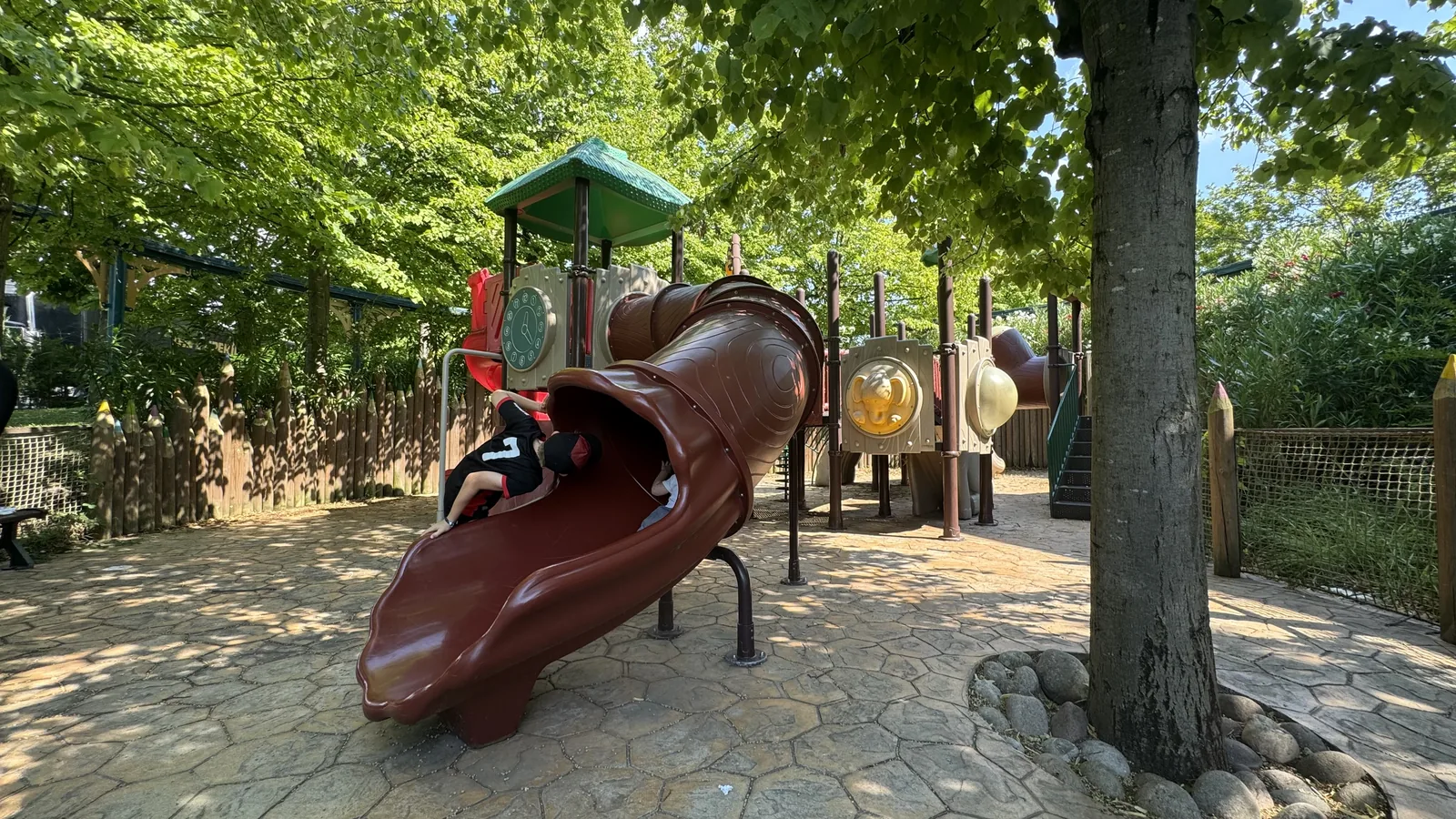 Schattiger Spielplatz mit brauner Röhrenrutsche und hölzernem Kletterturm unter hohen Bäumen im Movieland Park, ein Kind steigt in die Rutsche