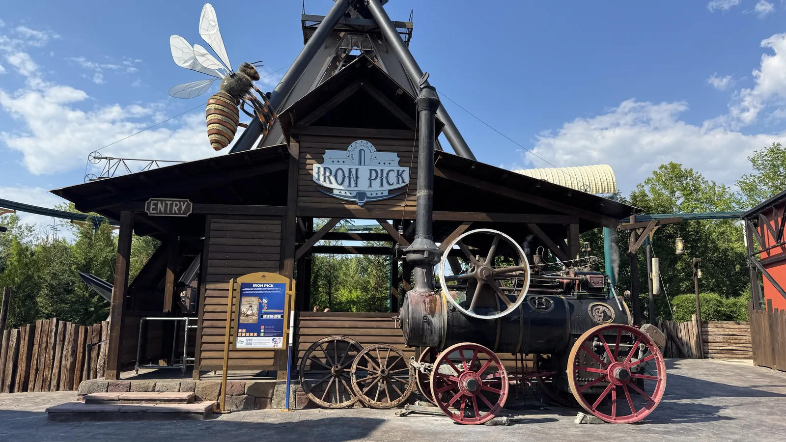 Dunkles Holzgebäude mit den Aufschriften Iron Pick und Entry, davor eine alte Dampfmaschine mit roten Speichenrädern, auf dem Dach eine überlebensgroße Bienenfigur