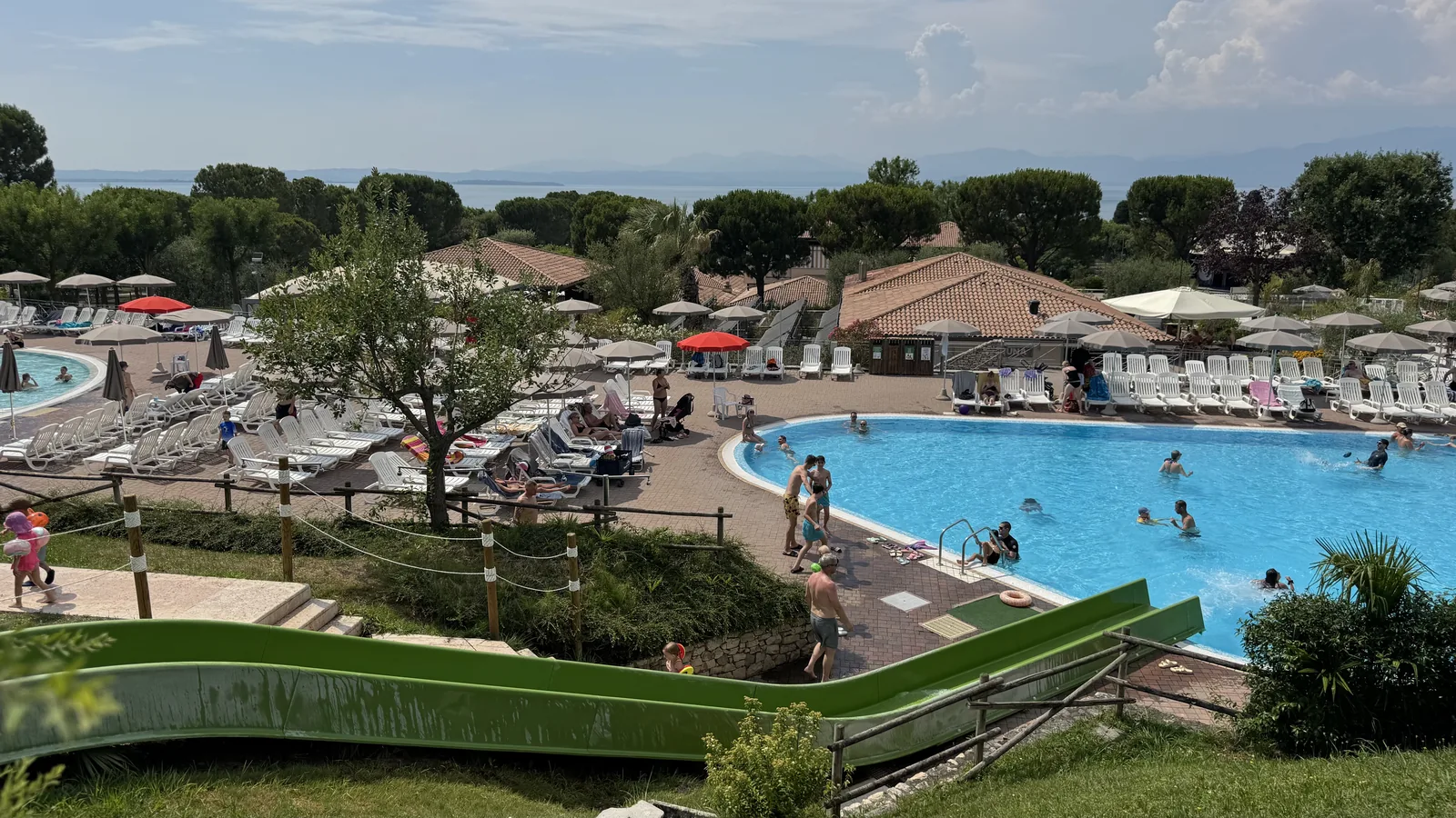 Blick über die Poollandschaft von Camping Piani di Clodia mit grüner Rutsche, großem Becken und dem Gardasee im Hintergrund