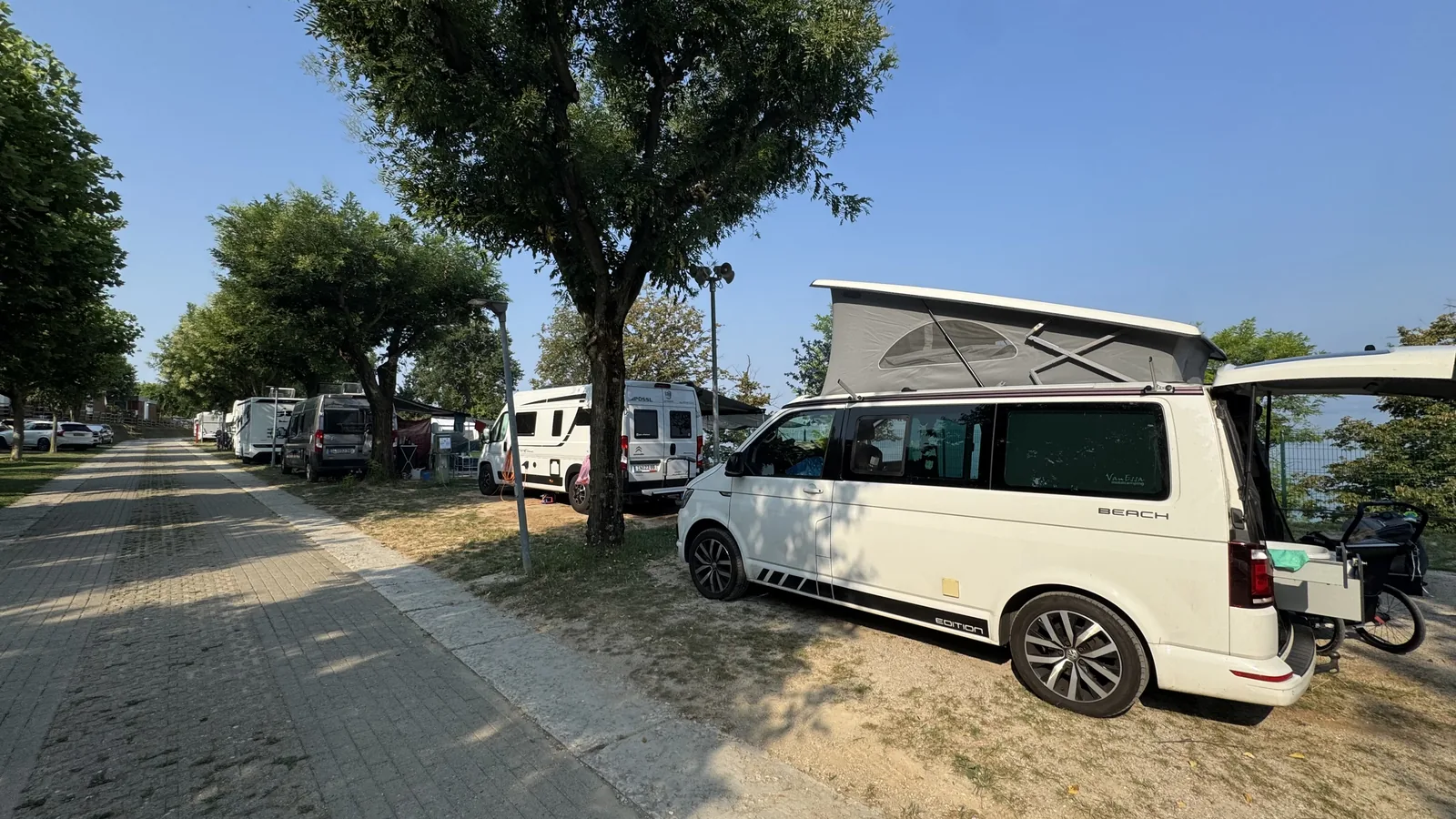 Weißer VW California mit aufgestelltem Hubdach unter Laubbäumen auf dem Campingplatz Piani di Clodia, rechts der Gardasee