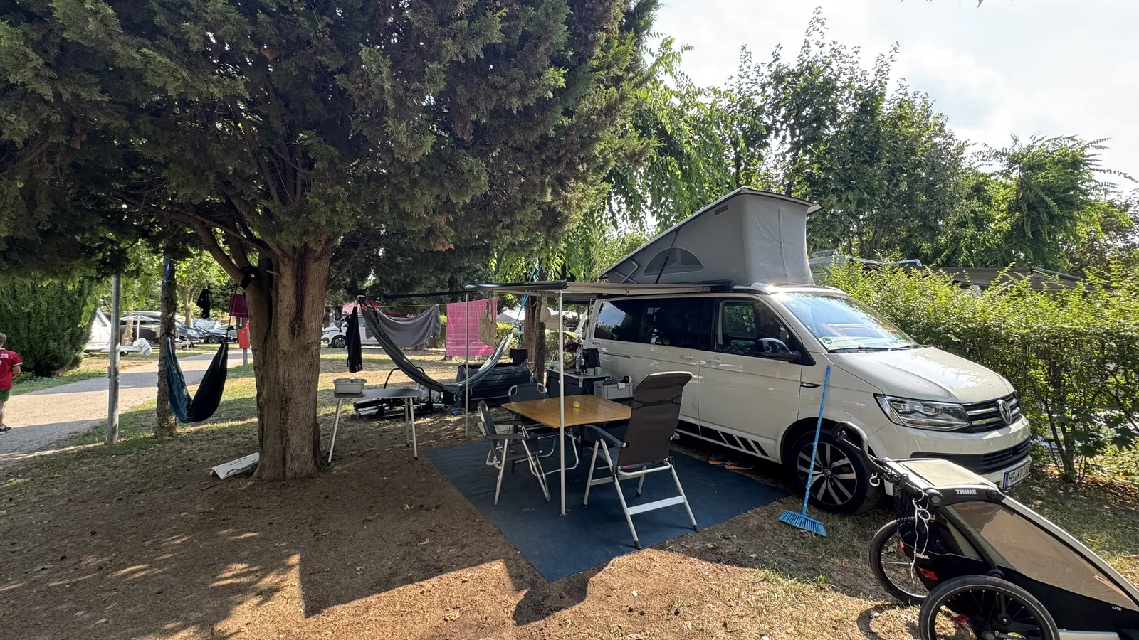 VW California mit ausgefahrener Markise, Campingtisch, vier Stühlen und zwei Hängematten auf einem parzellierten Stellplatz