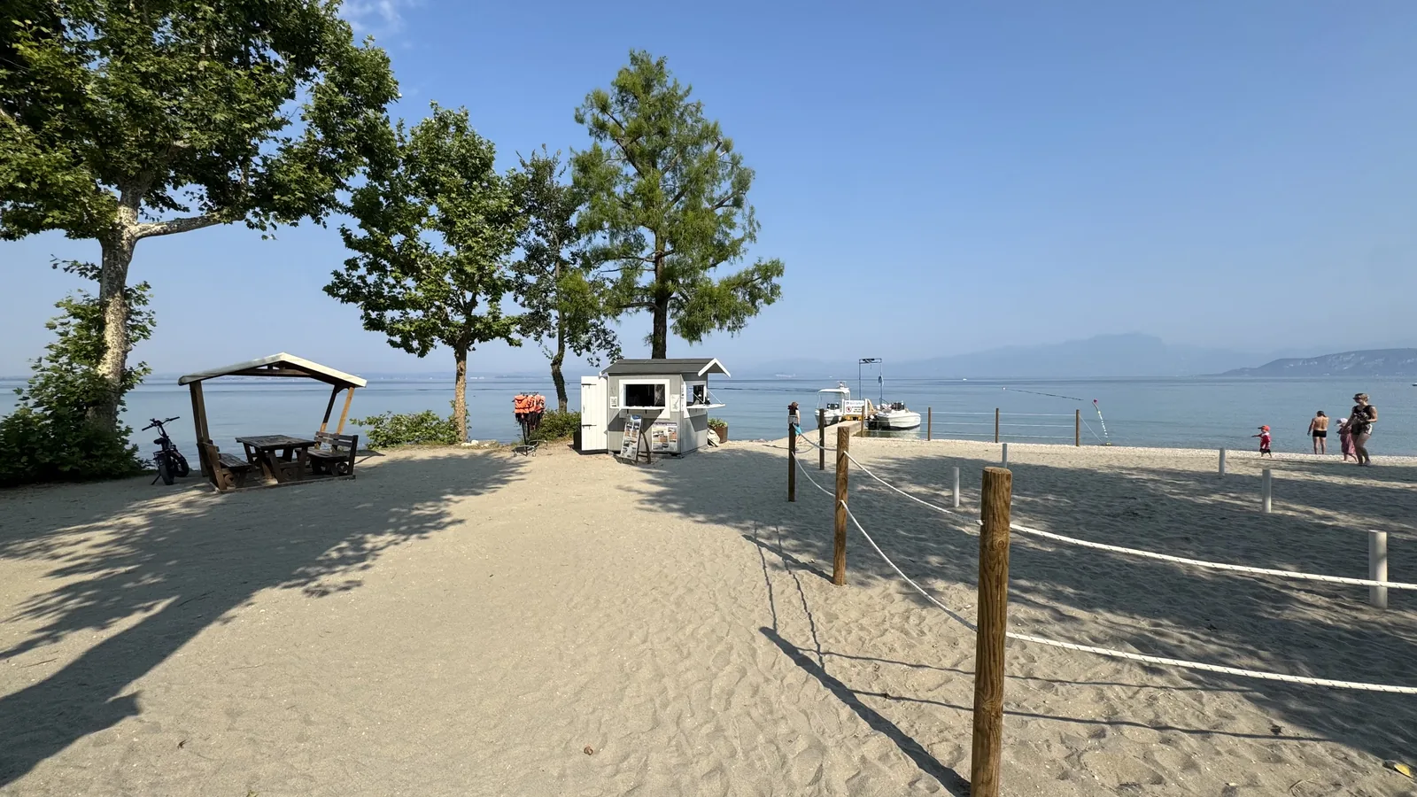 Aufgeschütteter Sandstrand am Gardasee mit Schattenbäumen, Picknickplatz, Kiosk, Steg mit Leihbooten und Bergpanorama