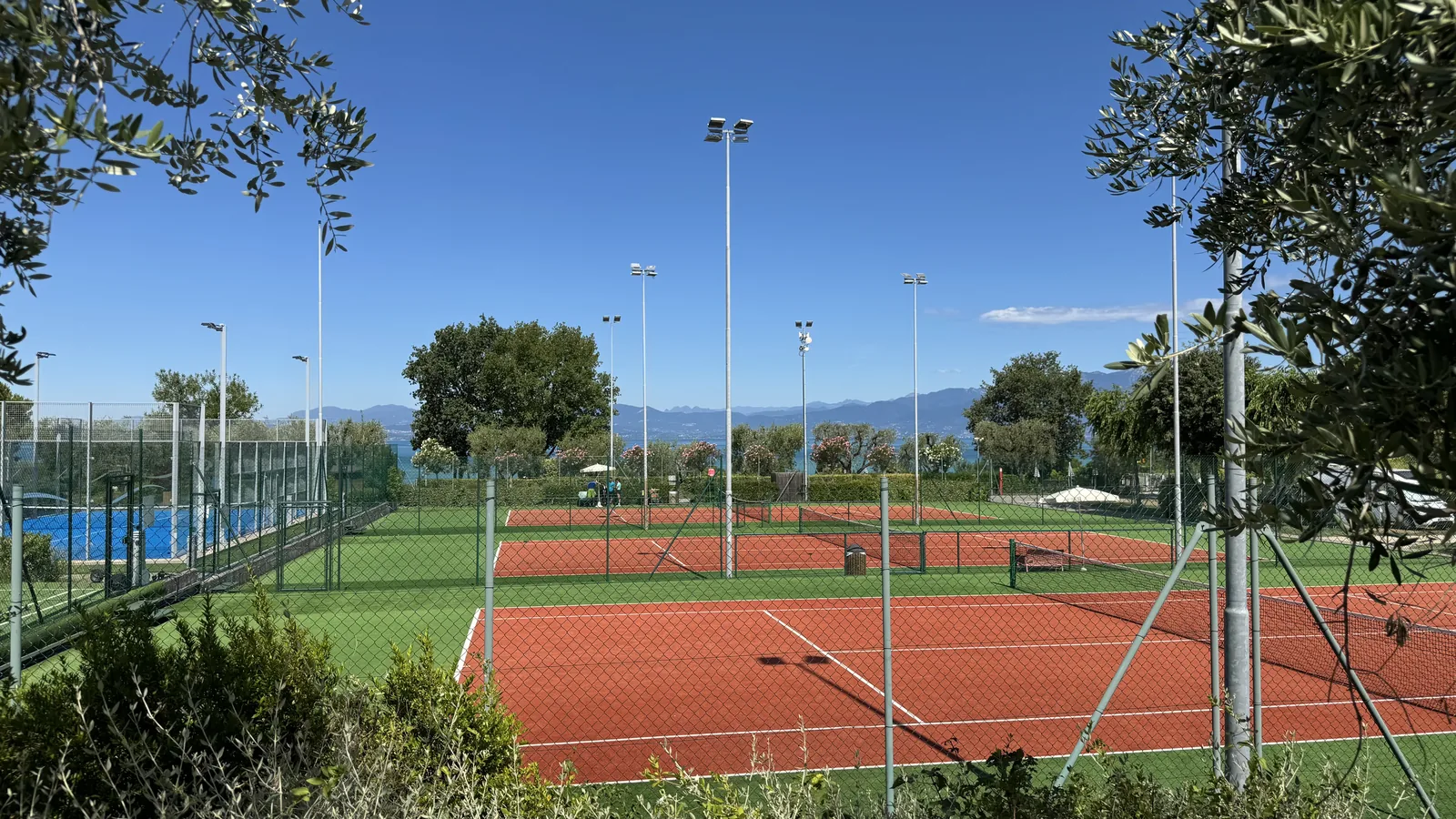 Sandtennisplätze mit Flutlichtmasten und blauem Padelplatz auf Camping Piani di Clodia, dahinter der Gardasee und die Berge