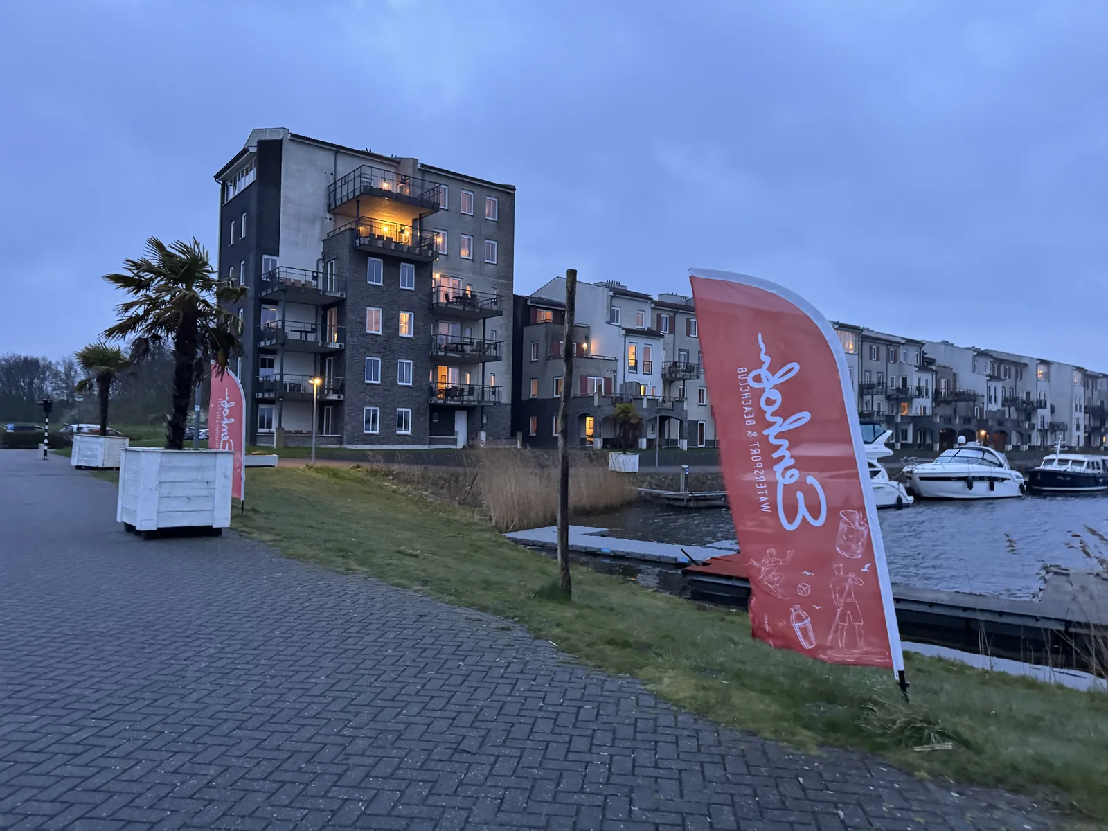 Uferpromenade in der Dämmerung mit roter Beachflag, Apartmenthäusern und Booten im Hafen