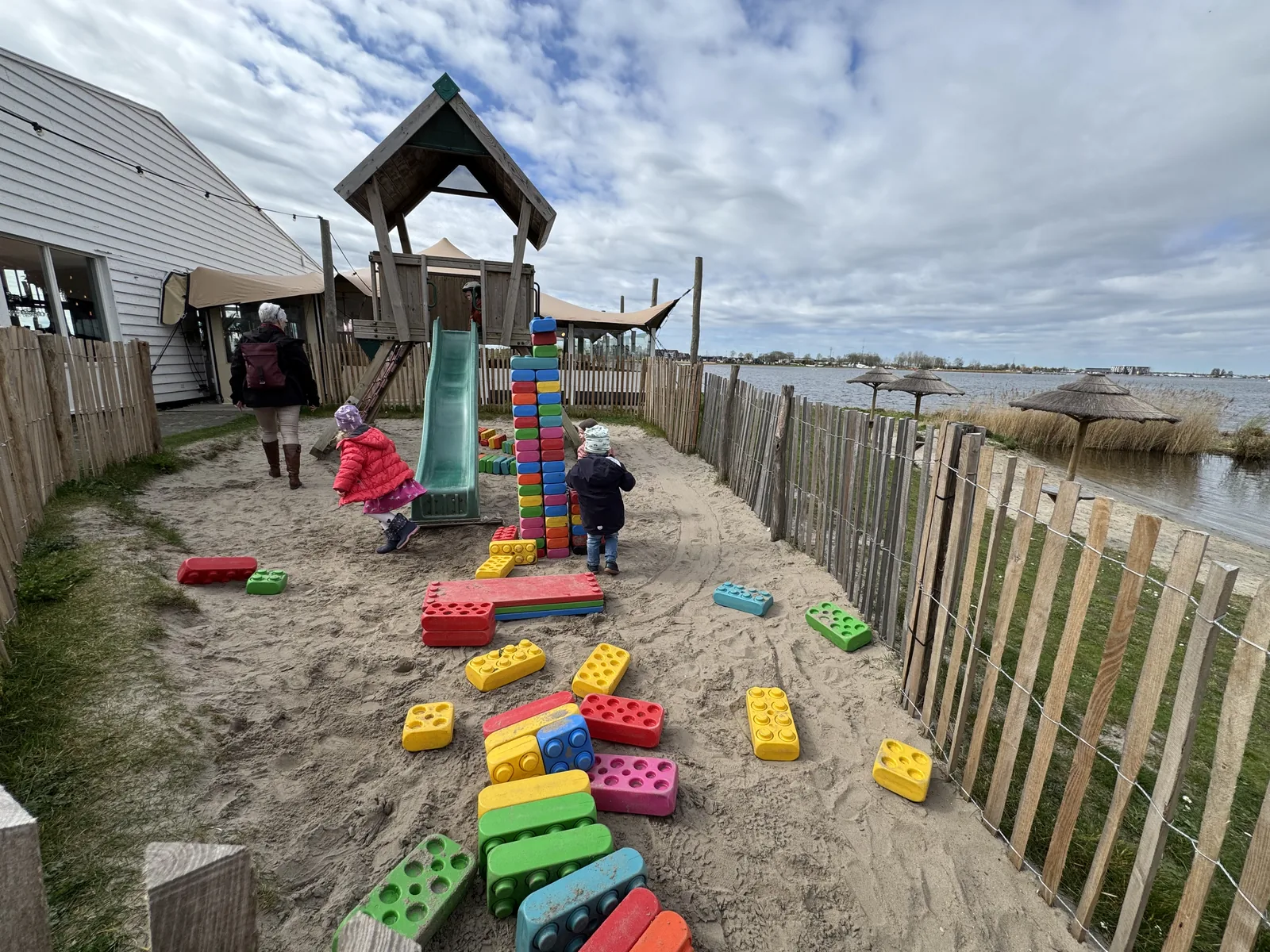 Sandspielplatz mit Holzkletterturm, Rutsche und großen bunten Bausteinen hinter einem Lattenzaun am Wasser