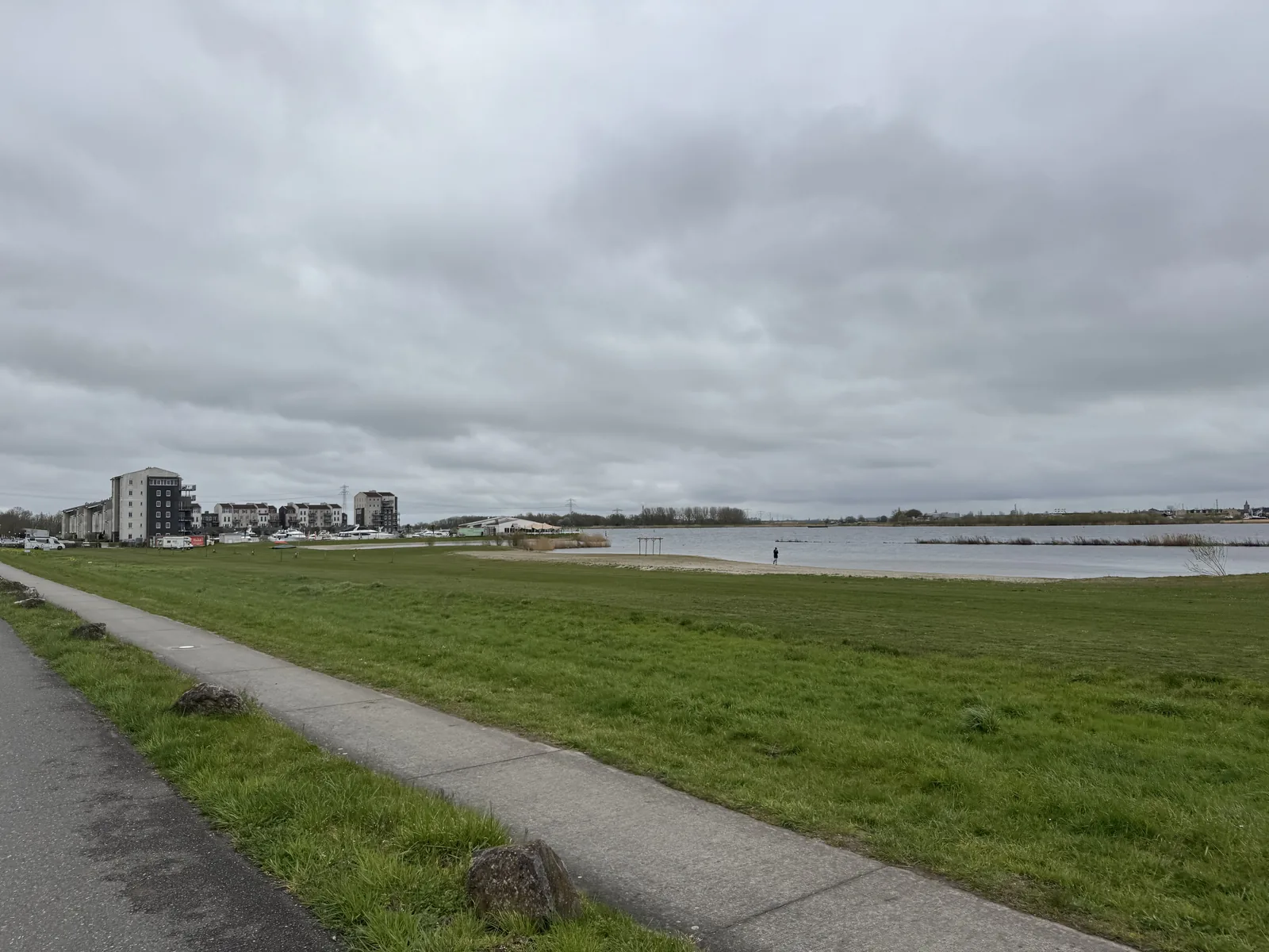 Asphaltierter Uferweg zwischen Wiese und Eemmeer bei bedecktem Himmel, in der Ferne Gebäude am Wasser