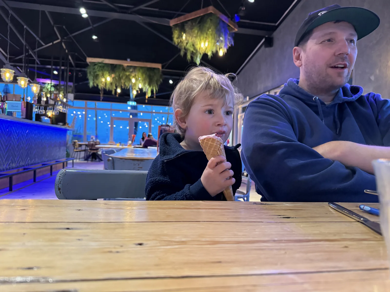 Mann mit Basecap und Junge mit Eiswaffel am Tisch im blau beleuchteten Beachclub