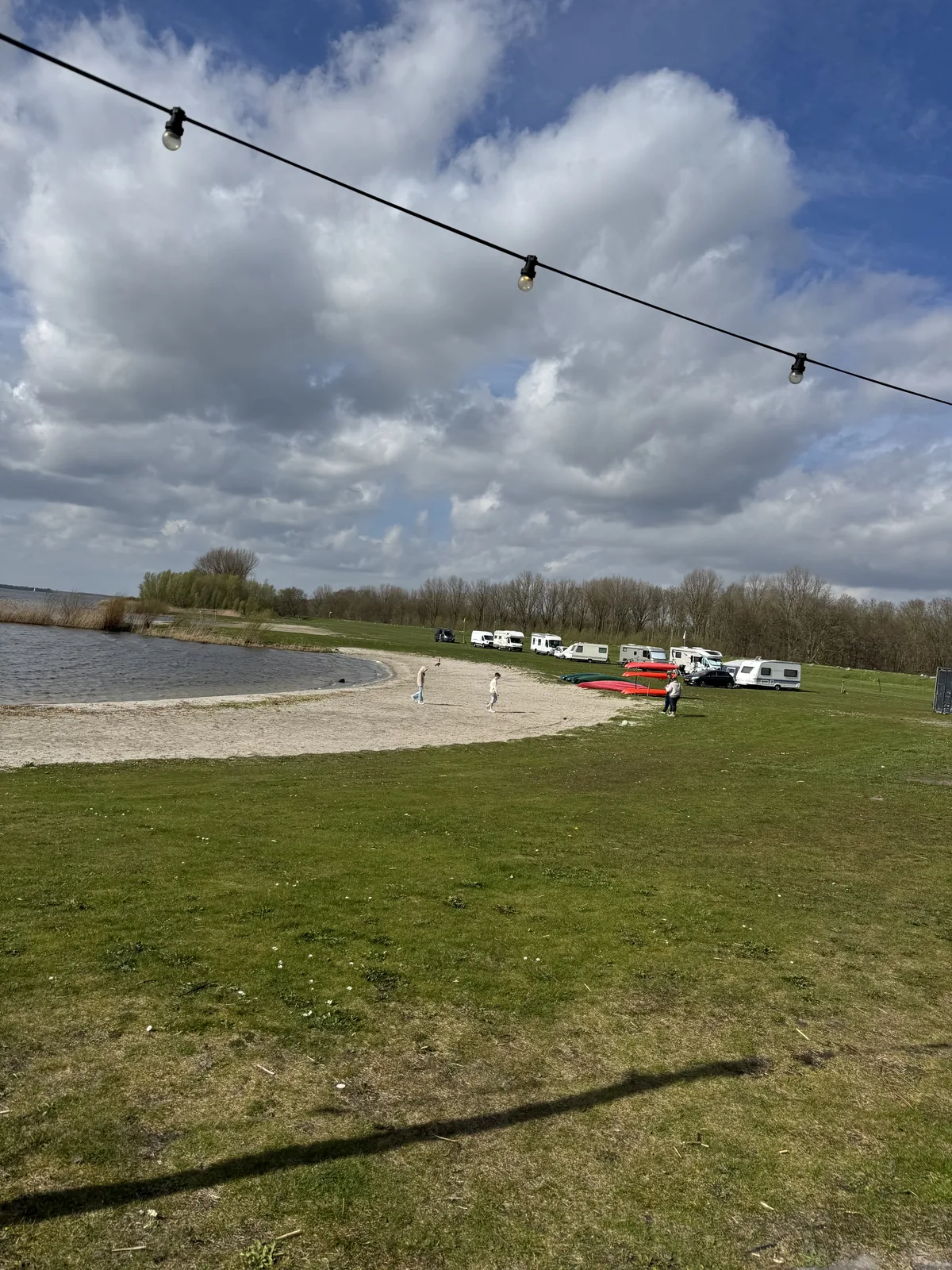 Weite Uferwiese mit gespannter Lichterkette, dahinter Wasser und Wolkenhimmel