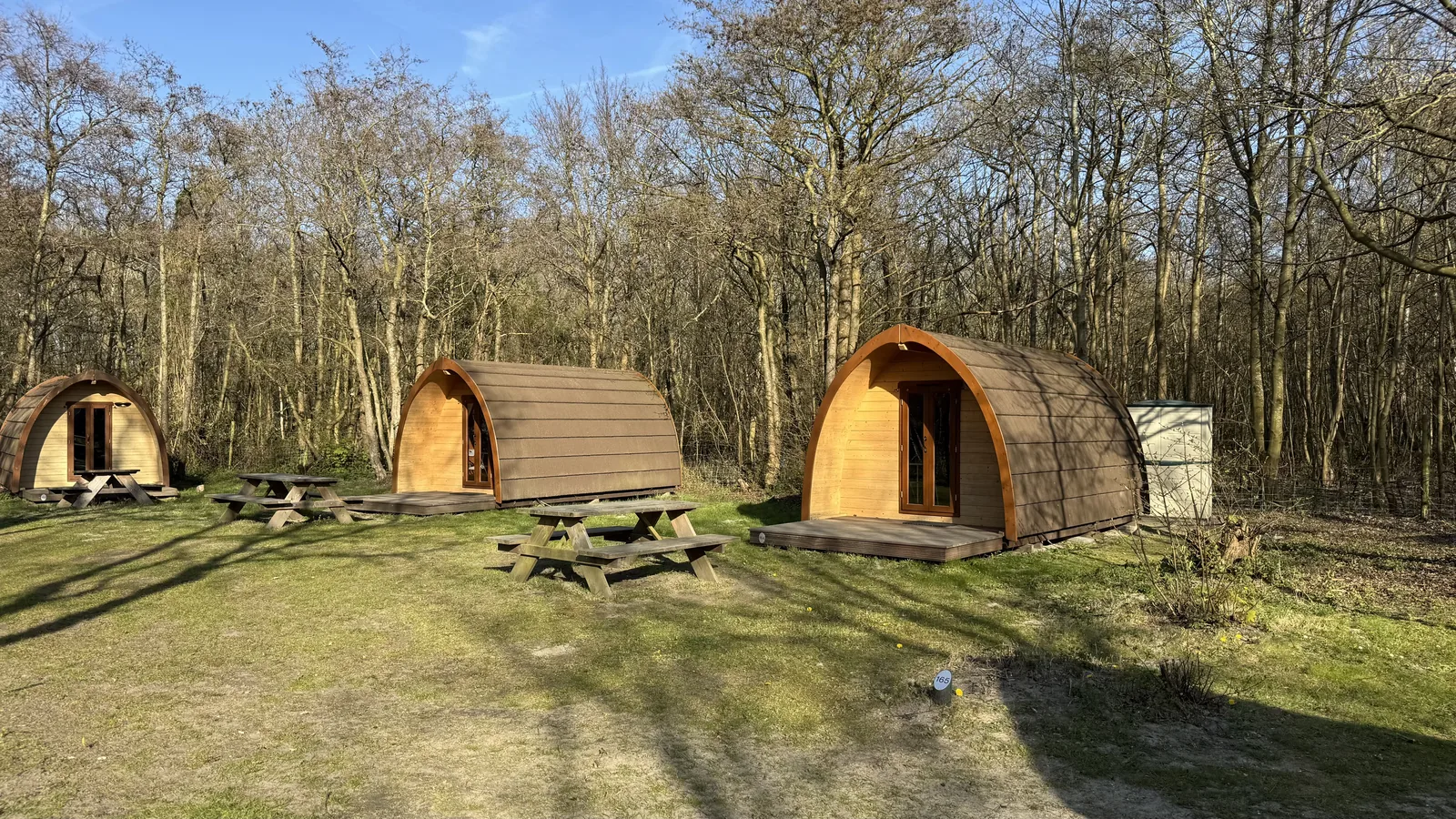 Zwei hölzerne Campingfässer mit Picknicktisch auf einer Wiese des Campingplatzes Port Zélande zwischen noch kahlen Bäumen