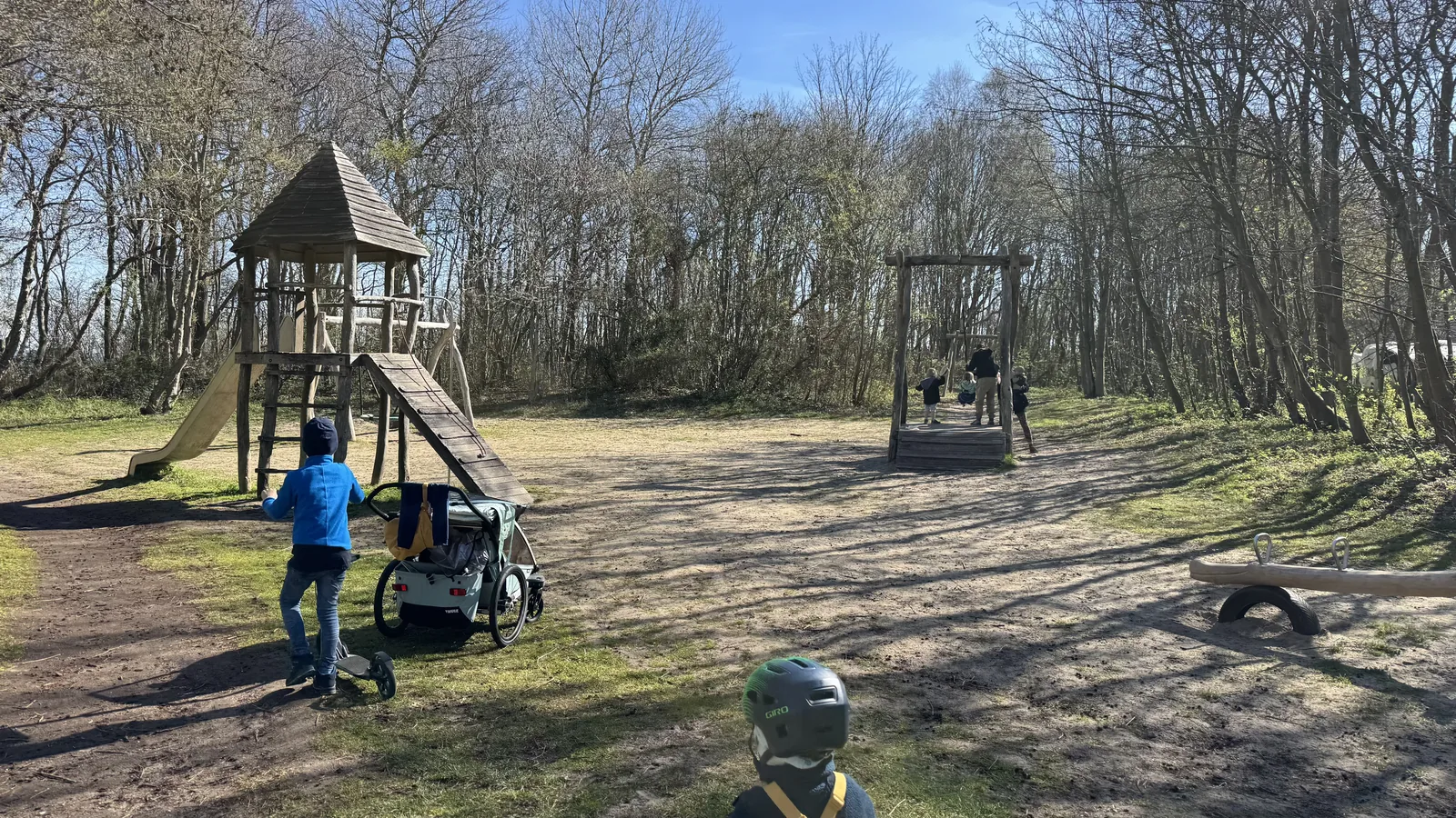 Naturnaher Spielplatz mit Holzturm, Rutsche und Reifenschaukel, im Vordergrund ein Kind mit Helm auf einem Roller