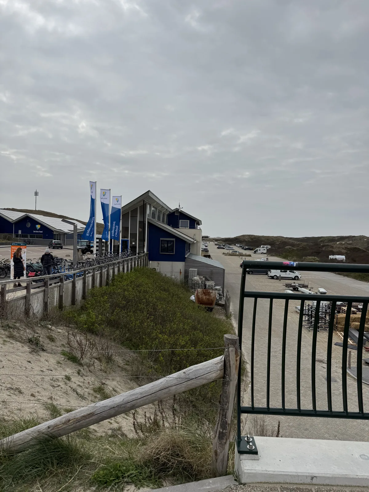 Dünenzugang mit Holzzaun und Treppengeländer, dahinter die blauen Gebäude von Camping Kogerstrand