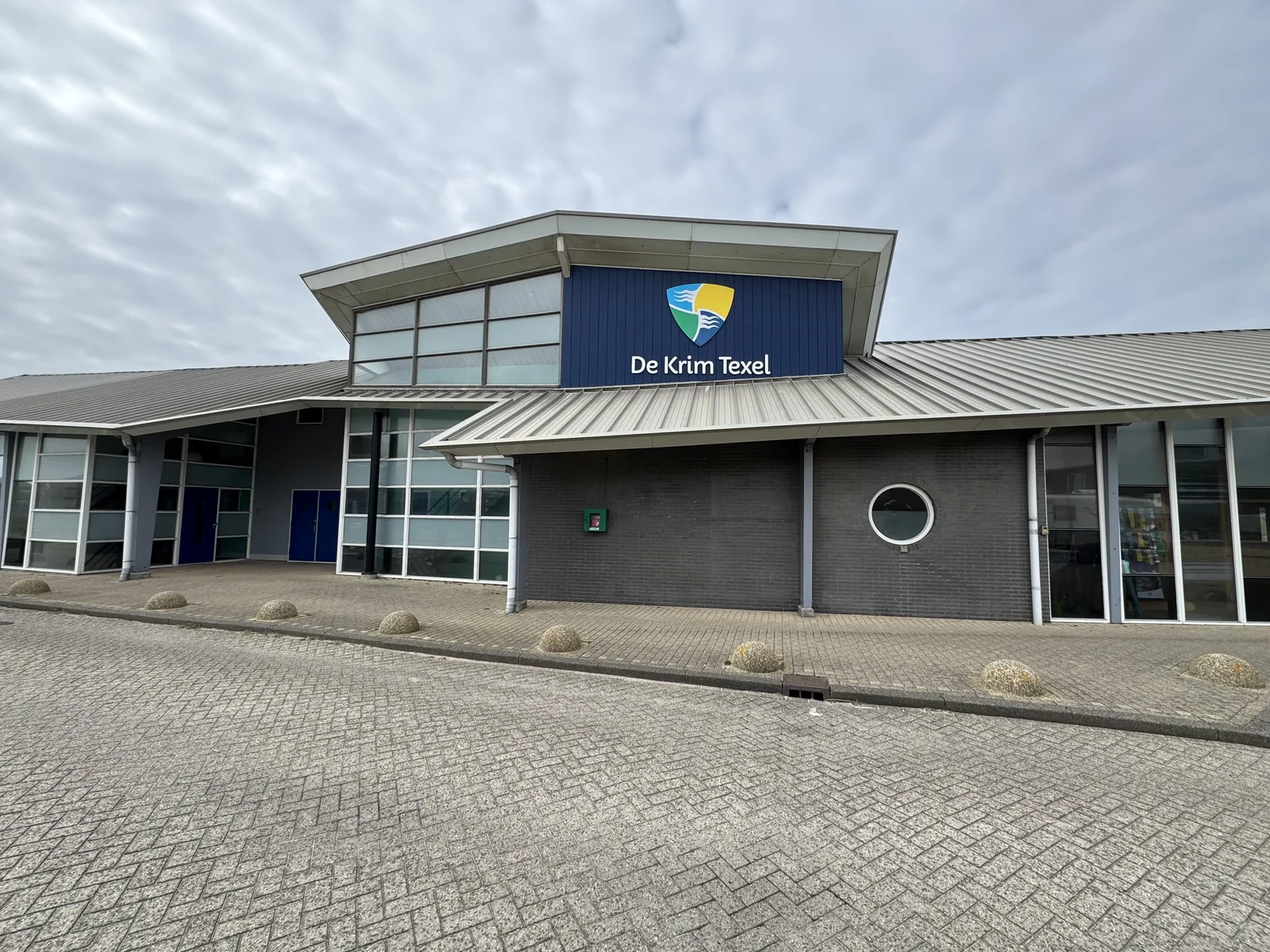 Hauptgebäude von Camping Kogerstrand auf Texel mit der Aufschrift De Krim Texel und großer Glasfront, davor der gepflasterte Vorplatz