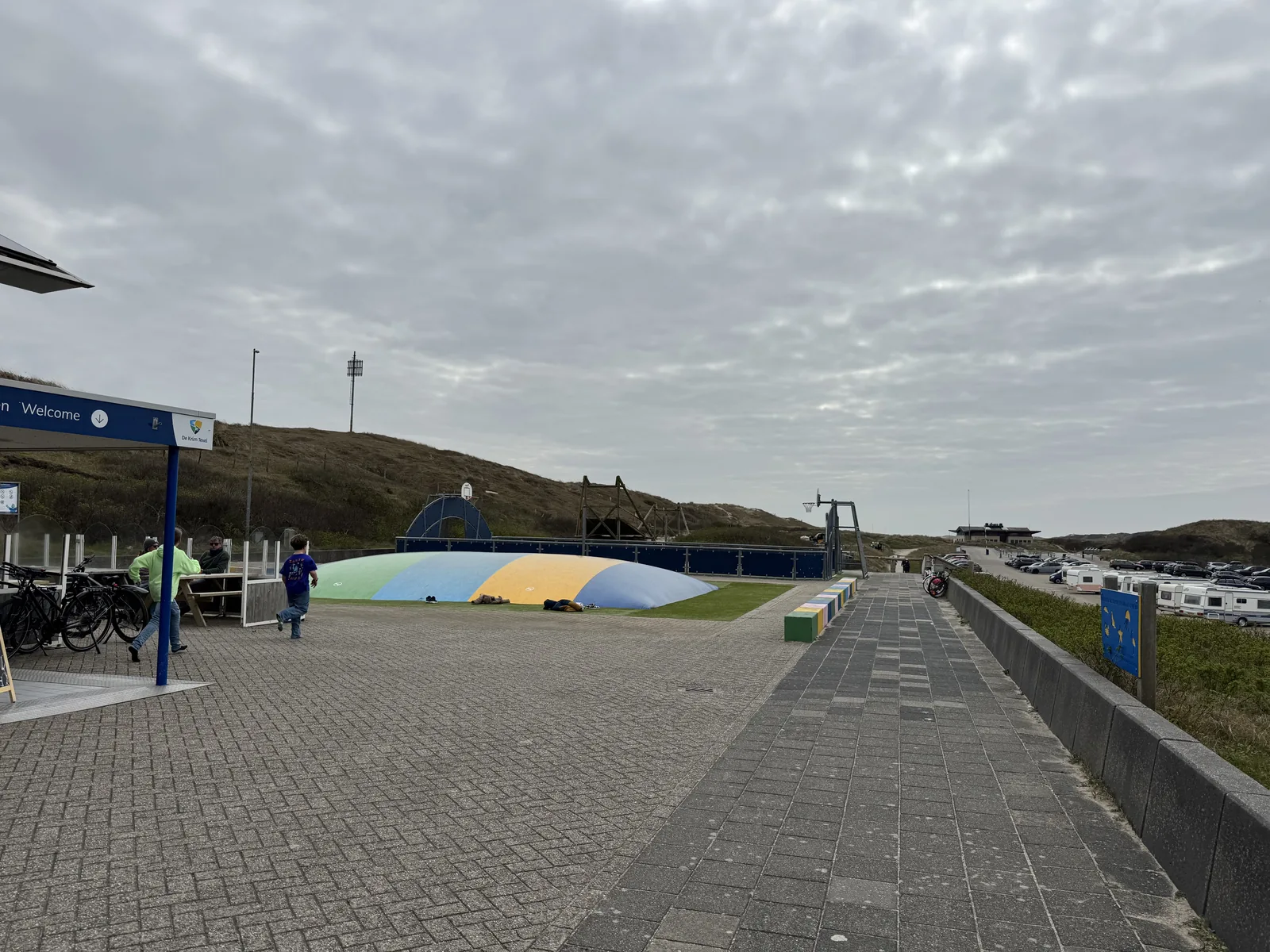 Breiter gepflasterter Weg am Eingang von Camping Kogerstrand mit farbigem Spielhügel, Fahrradständern und Dünenkulisse