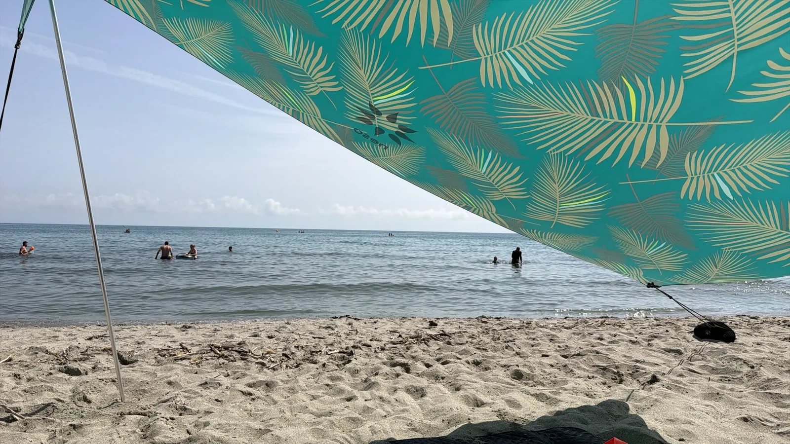 Blick unter dem Sonnensegel auf den schmalen Sandstreifen und das flache Wasser der Bucht 2026