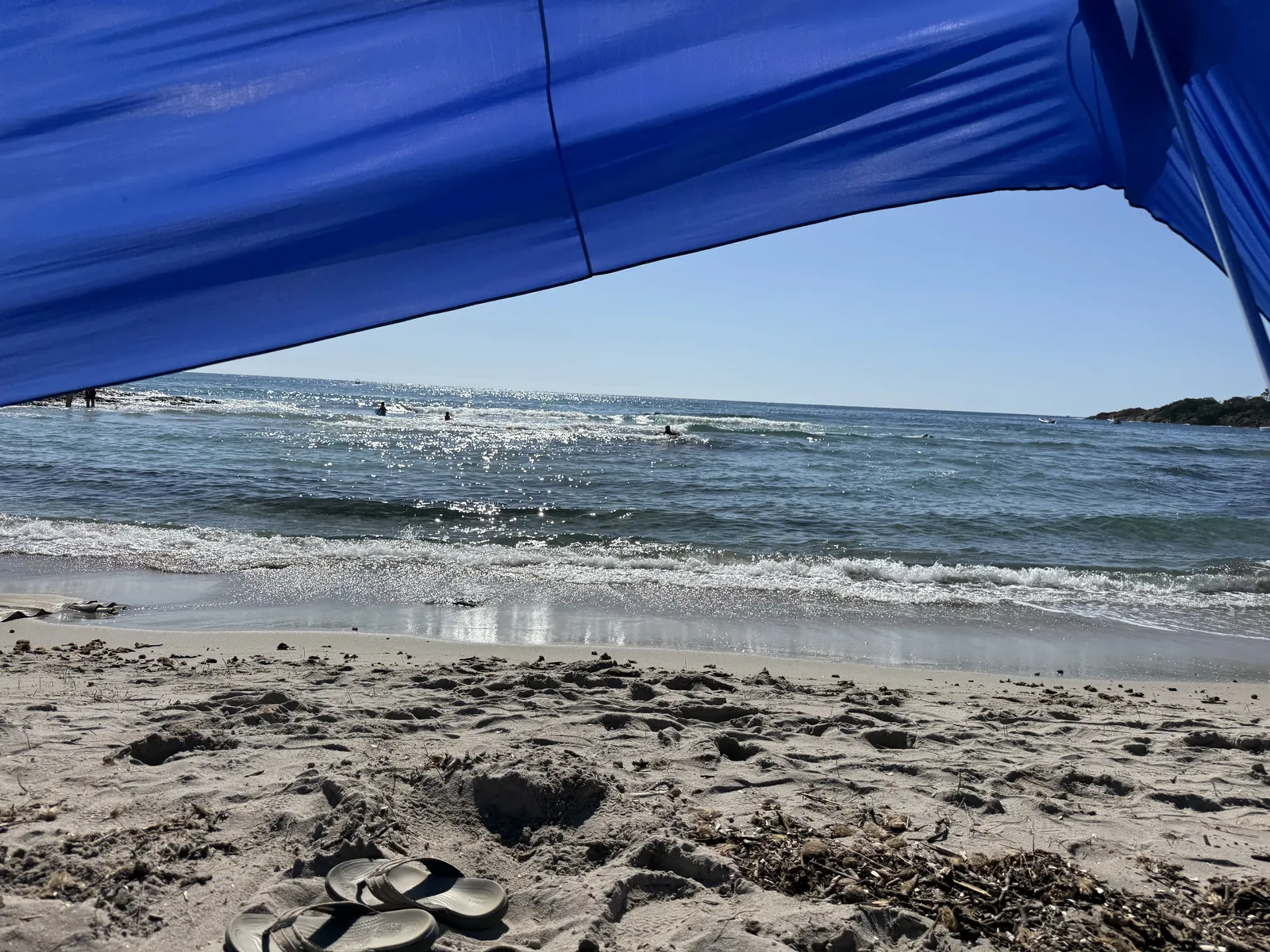 Blick unter dem Sonnensegel auf den flachen Strand und das ruhige Wasser der Bucht von Pinarello