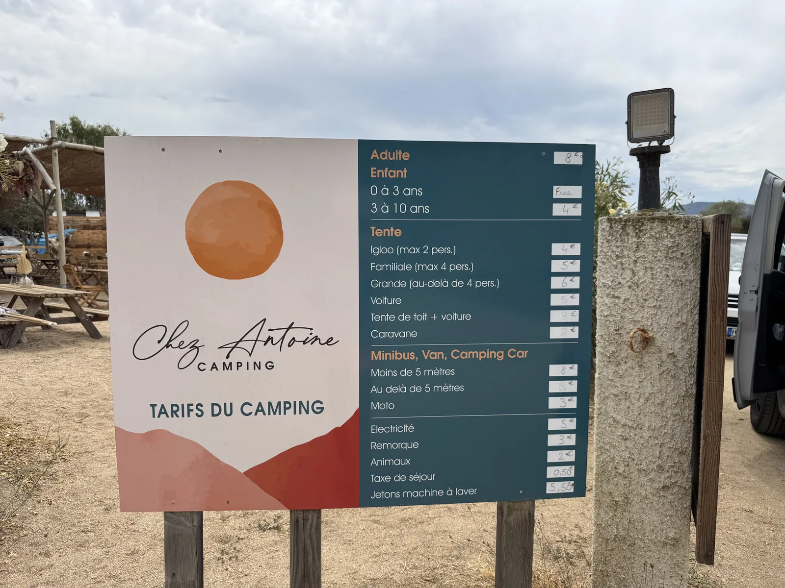 Eingangsschild mit Preistafel des Camping Chez Antoine, dahinter Picknicktische unter Olivenbäumen