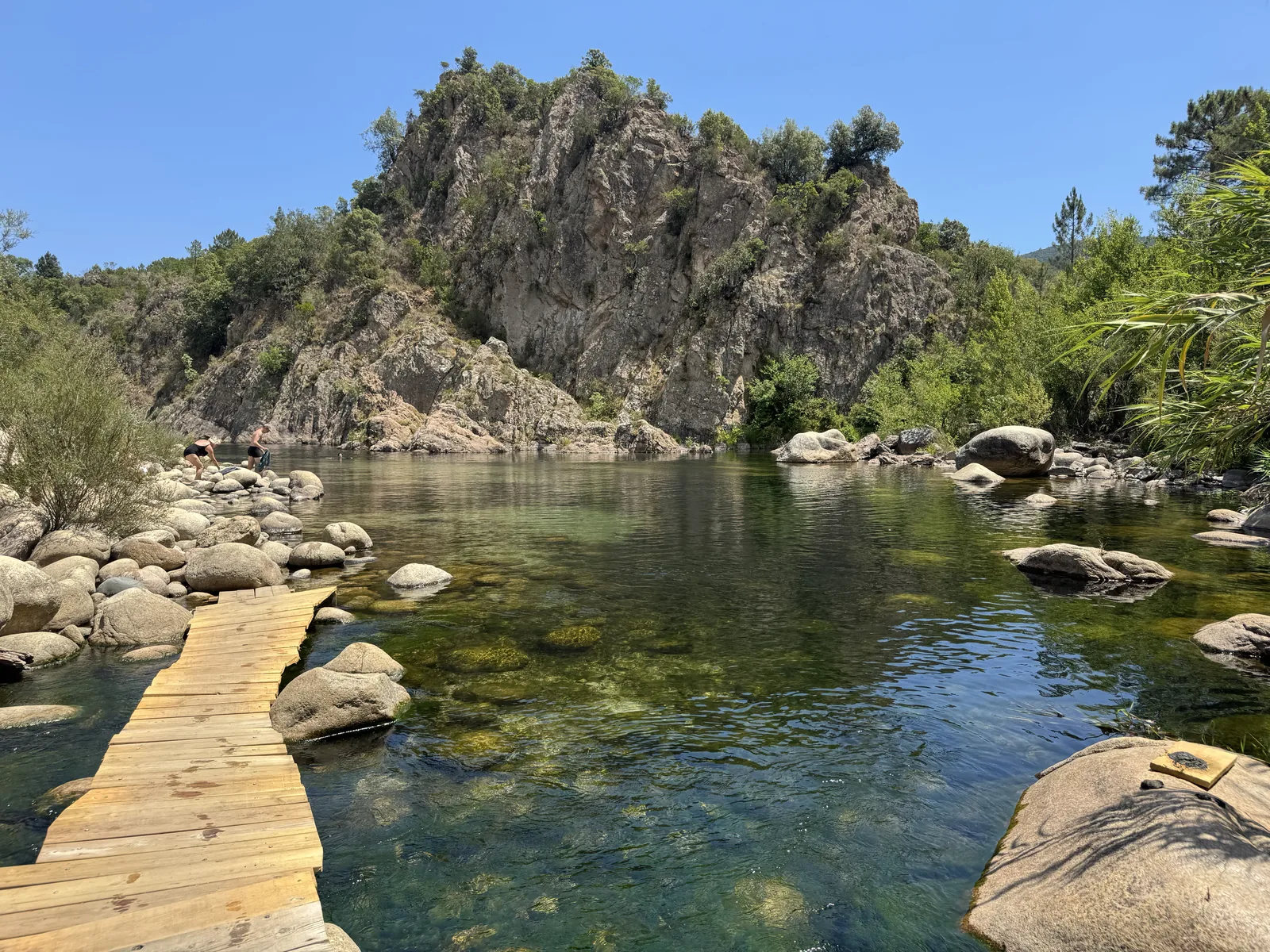 Holzsteg über den klaren Fluss vor einem Felsmassiv am Camping U Rosumarinu