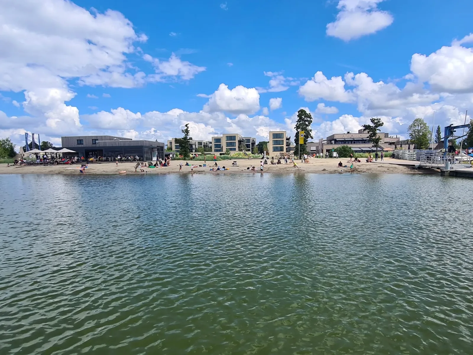Blick über den Freizeitsee auf das Strandbad des Marissa Ferienparks mit Sandstrand, Beachbar und den Apartmenthäusern dahinter