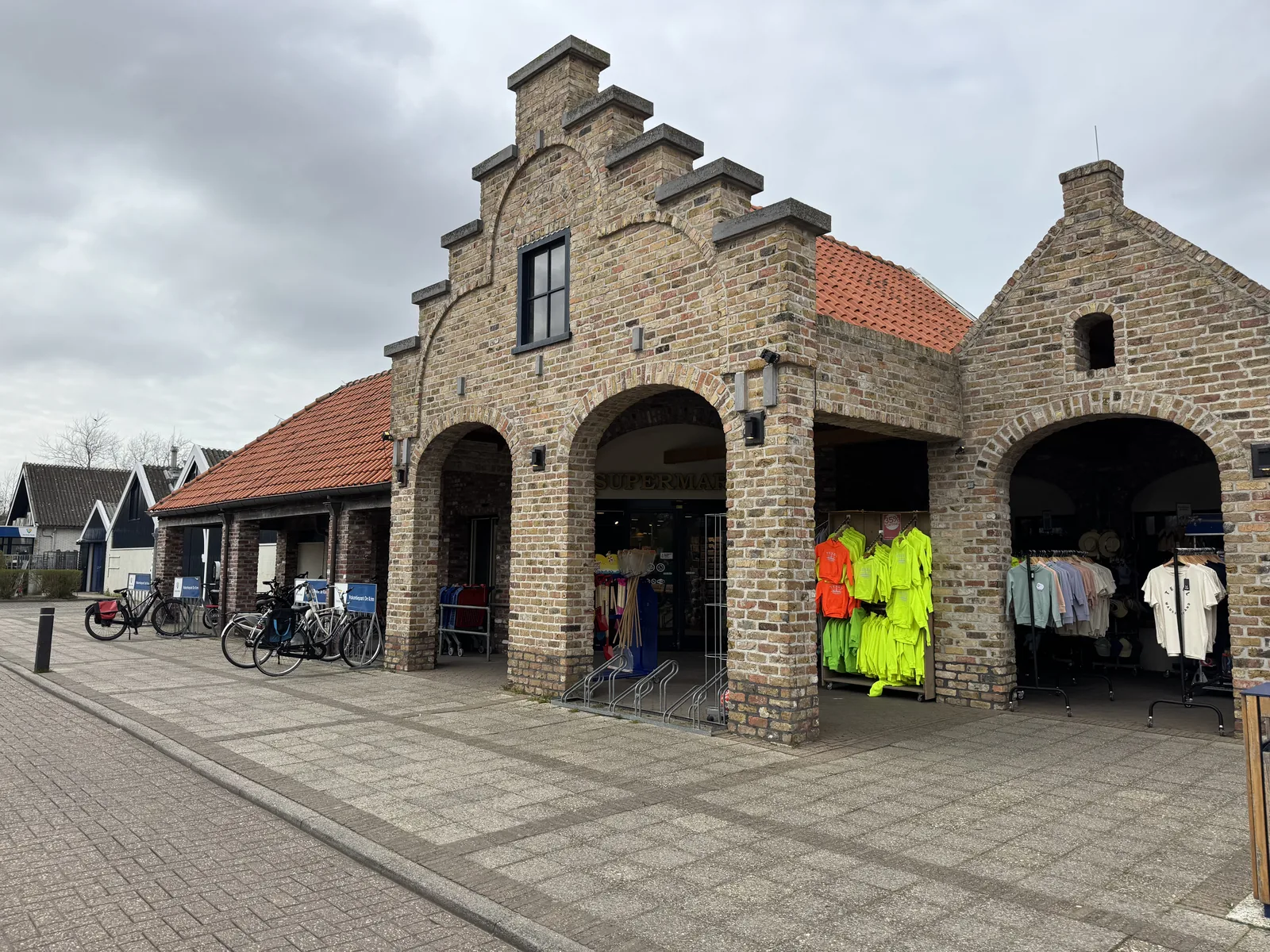 Backsteingebäude mit Staffelgiebel und Torbögen im Ferienpark De Krim, darunter Läden mit ausgehängter Kleidung und abgestellte Fahrräder
