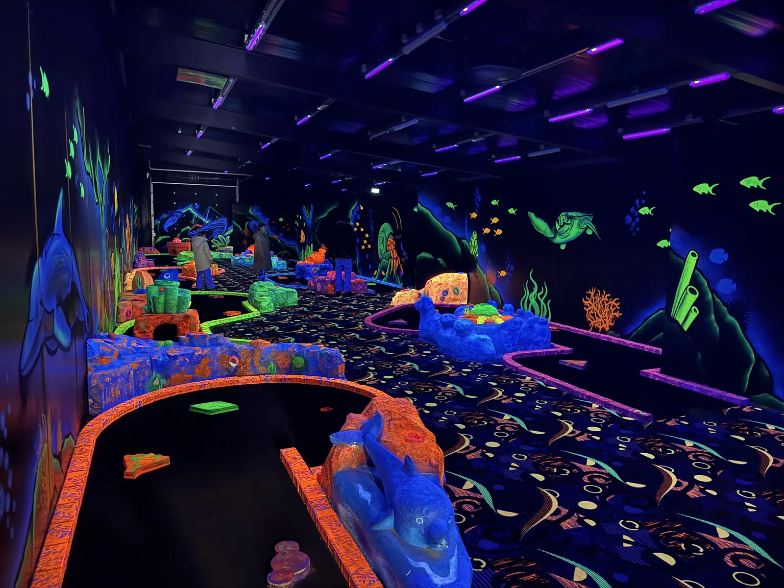 Schwarzlicht-Minigolf im Unterwasser-Look mit leuchtenden Fischen, Korallen und Bahnen im dunklen Raum