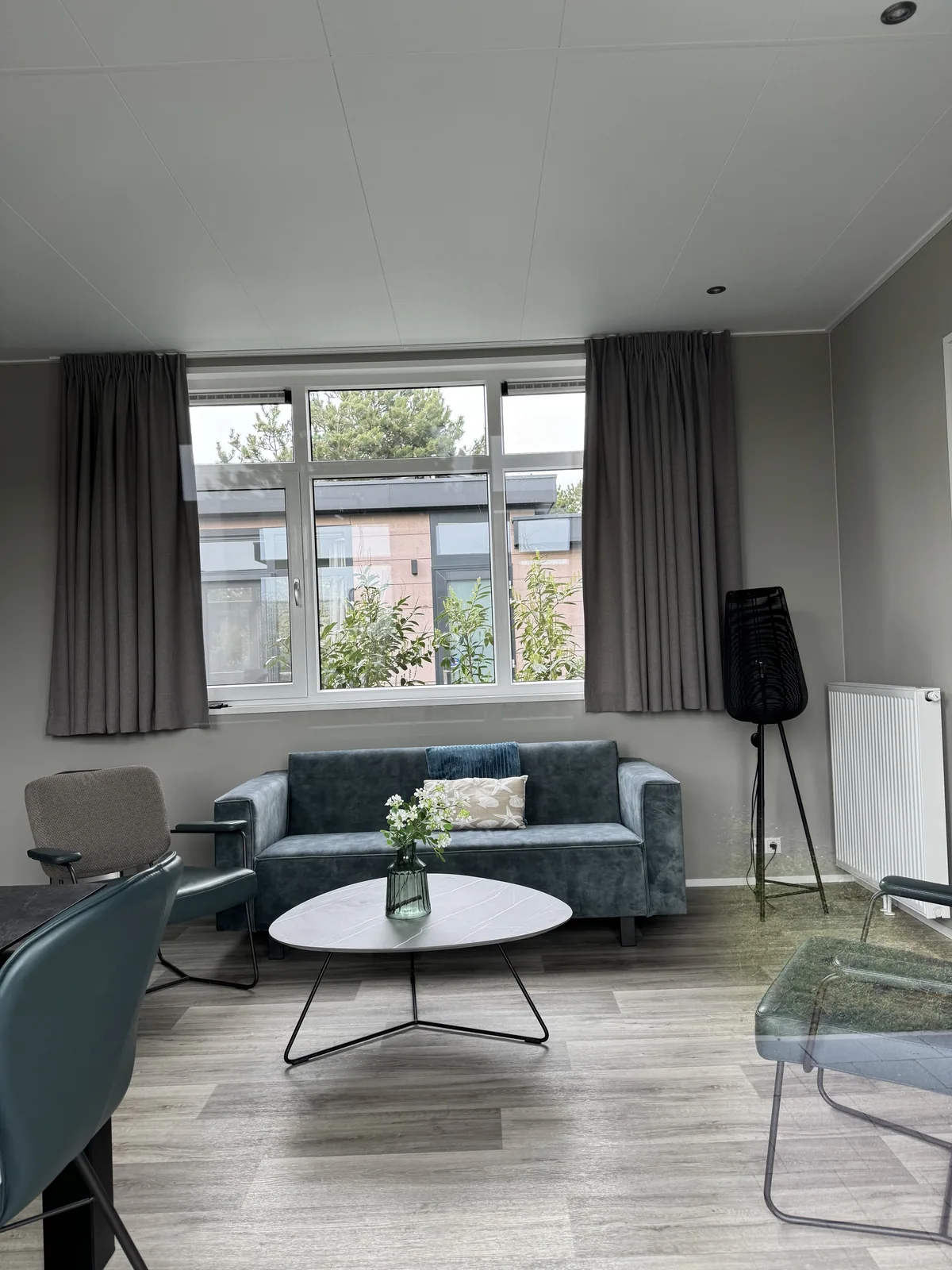 Wohnzimmer in einem Ferienhaus im Ferienpark De Krim mit petrolfarbenem Sofa, rundem Couchtisch und Fenster mit Vorhängen