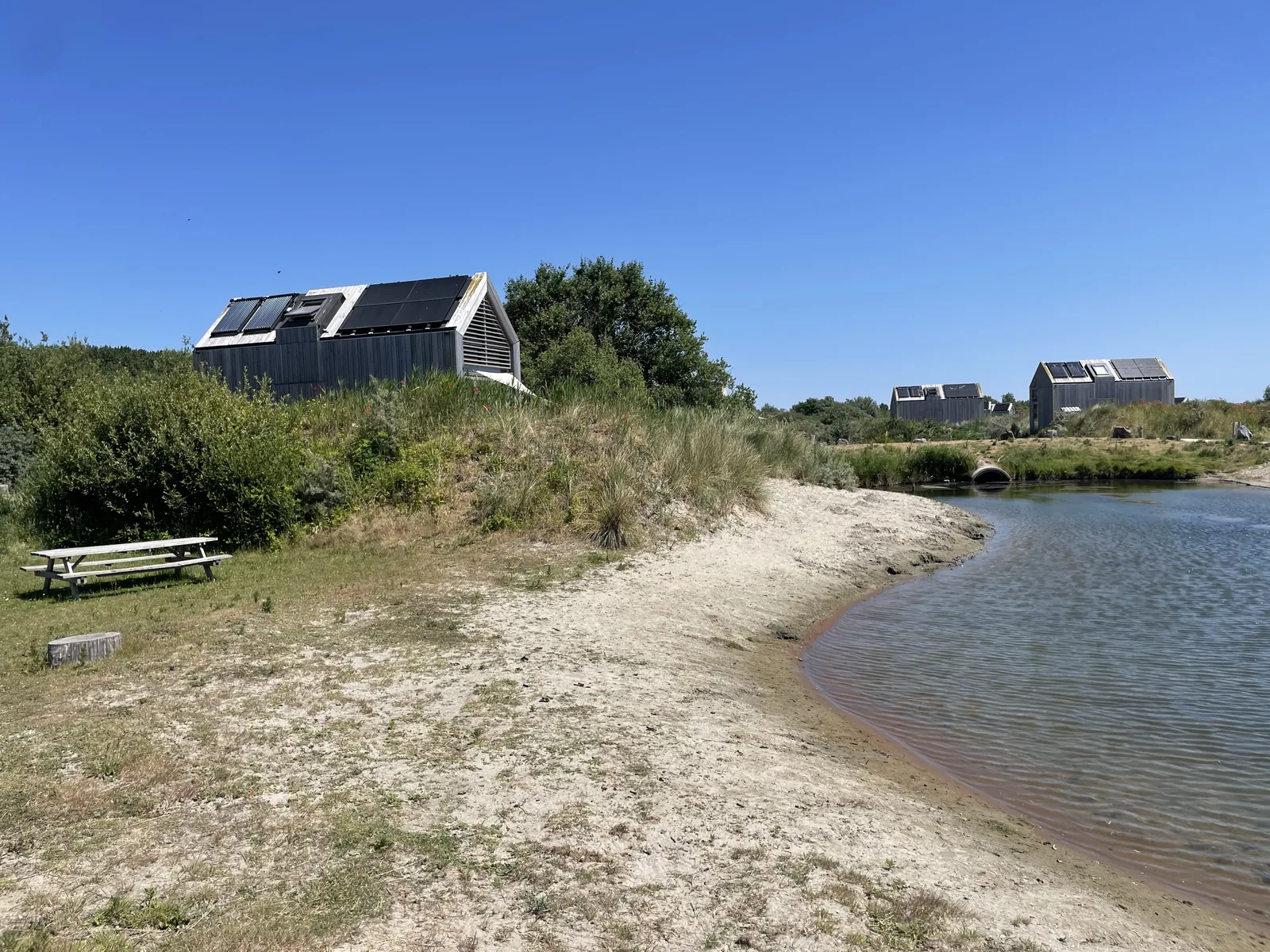 Zwei dunkle Ferienhäuser mit Solarpaneelen auf einer bewachsenen Düne, davor ein Sandufer und ein schmaler Wasserarm