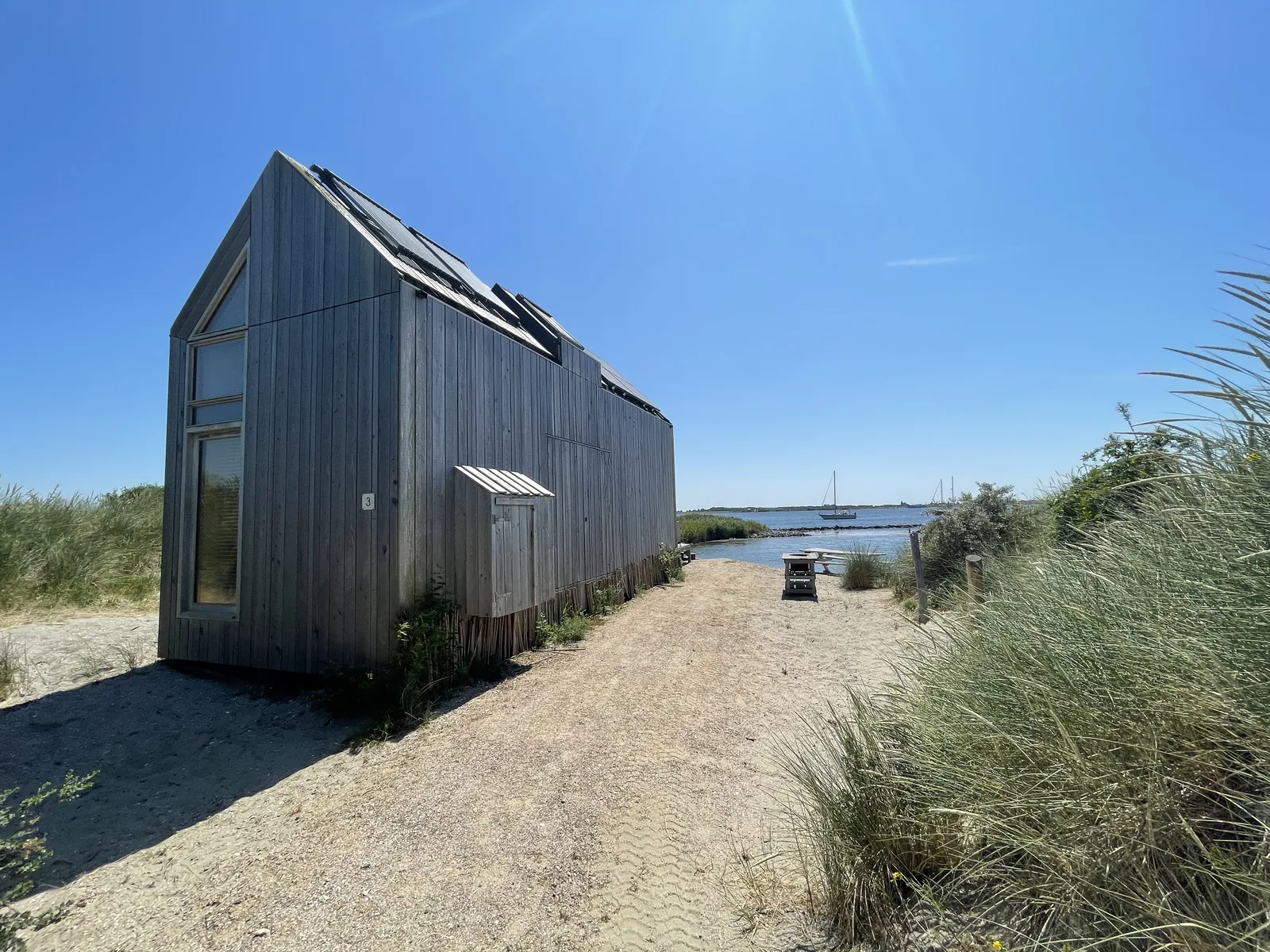 Seitenansicht eines schmalen, hellgrau verwitterten Holz-Ferienhauses mit hohem Giebel und Dachfenster, im Hintergrund Wasser und Segelboote