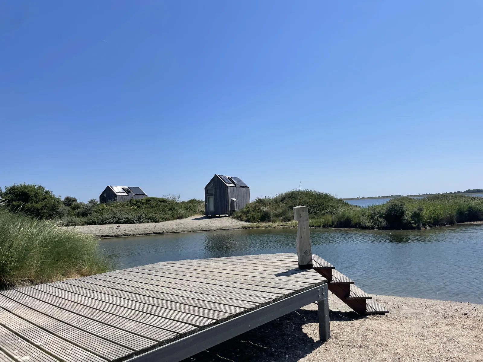 Hölzerner Steg mit Stufen ins flache Wasser der Bucht, gegenüber zwei dunkle Ferienhäuser auf der Landzunge