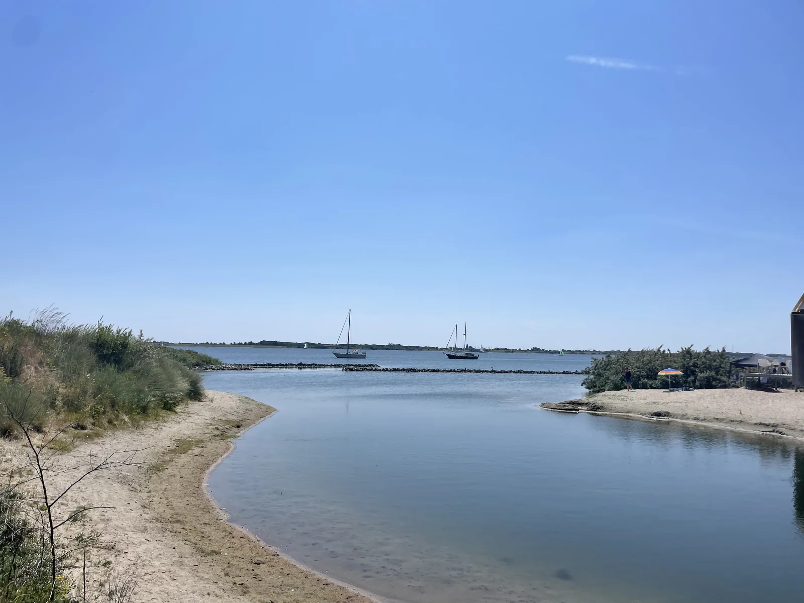 Sandiges Ufer mit Blick durch die Öffnung der Bucht auf das offene Grevelingenmeer, draußen zwei ankernde Segelboote