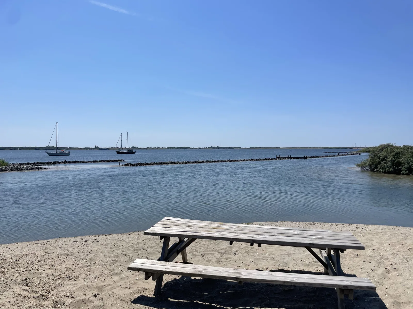 Verwitterte Picknickbank auf hellem Sand am Wasser, dahinter das Grevelingenmeer mit zwei Segelbooten und einem Steindamm