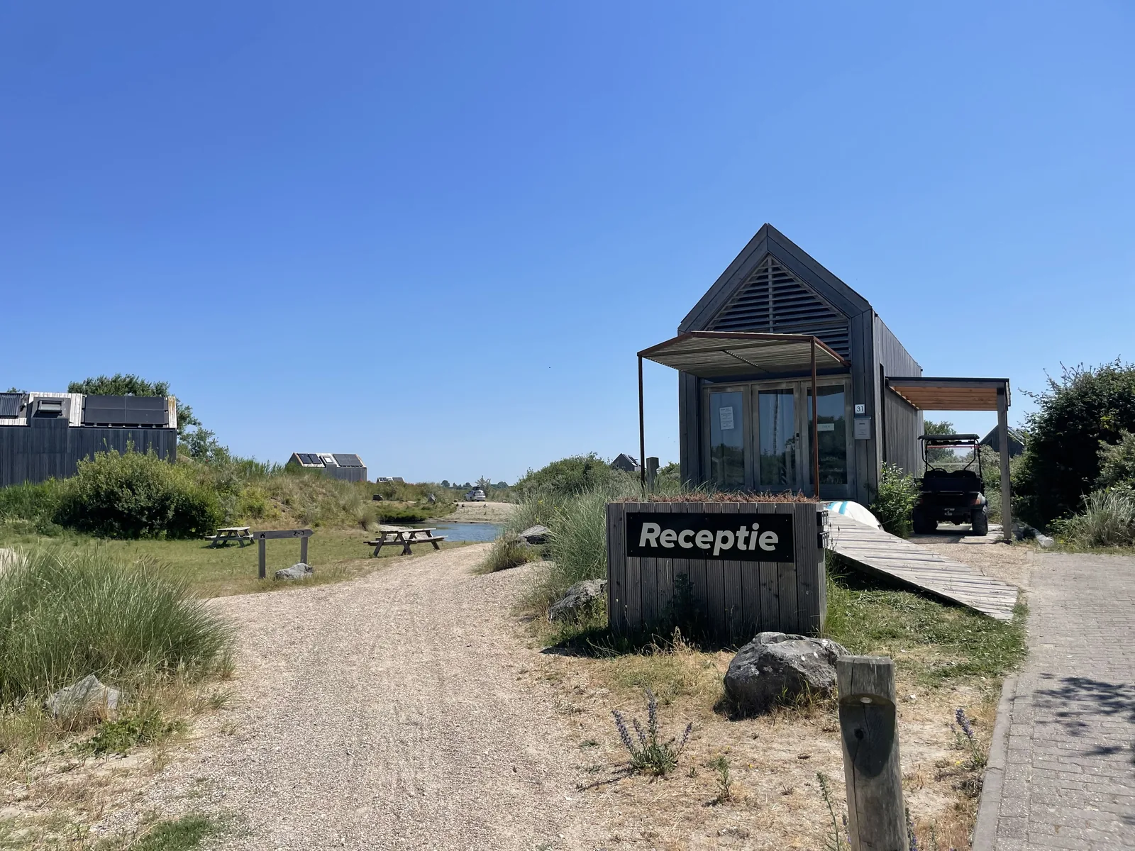 Kleines Rezeptionsgebäude aus Holz mit Vordach und schwarzem Schild Receptie, davor Picknickbänke auf der Wiese