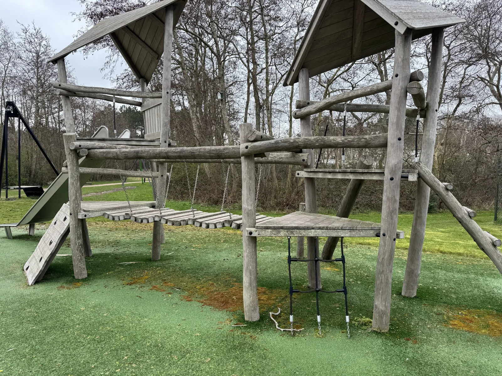 Großes hölzernes Spielgerüst mit zwei überdachten Türmen und Hängebrücke auf grünem Fallschutzbelag