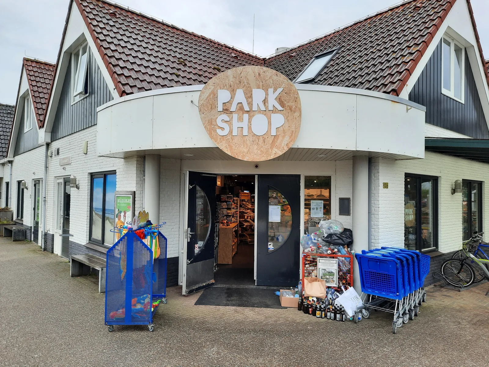 Außenansicht des Park Shop im Landal Sluftervallei mit rundem Holzschild, Einkaufskörben und Warenaufsteller vor der Tür