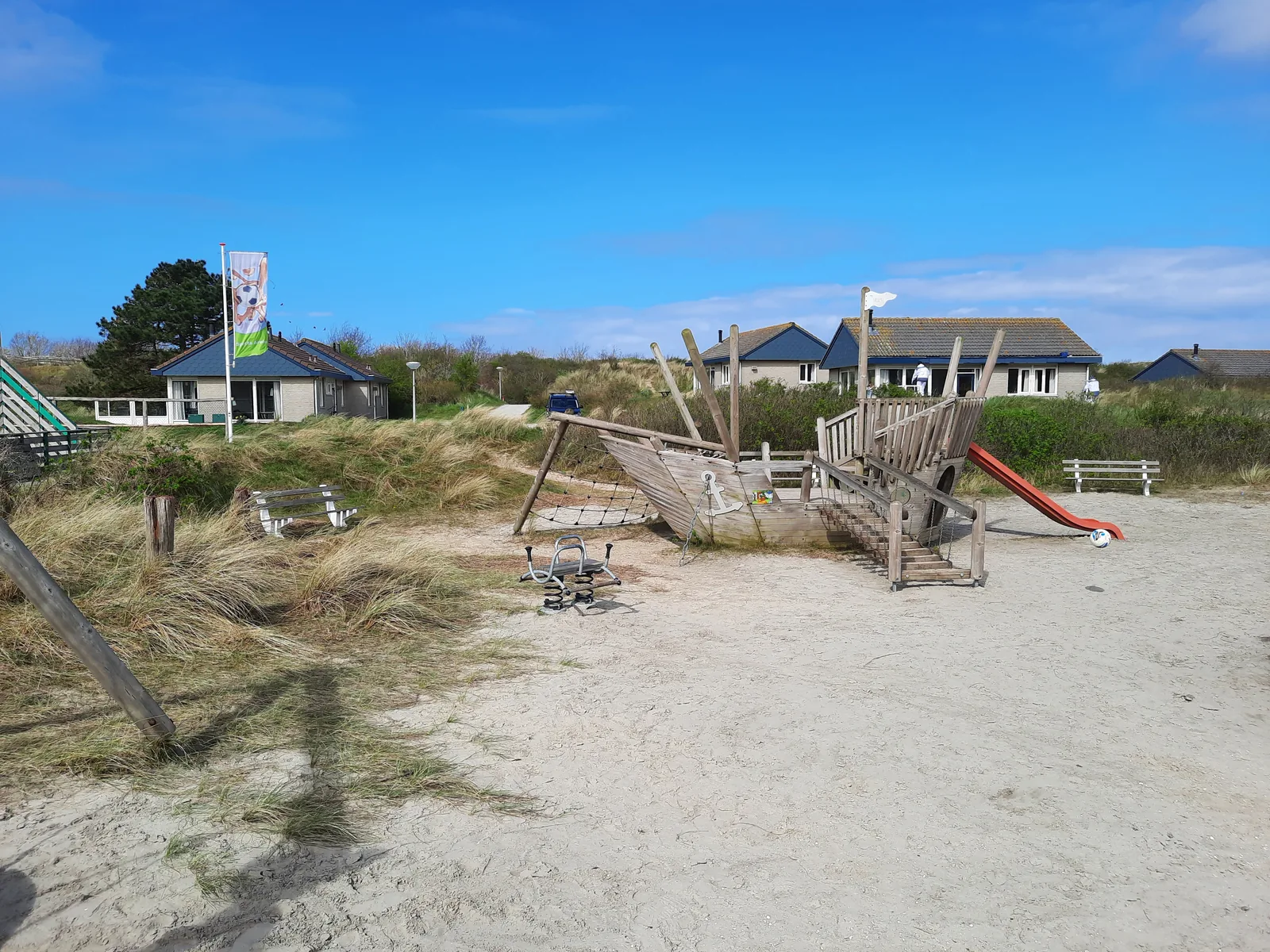 Piratenschiff-Spielgerät im hellen Sand am Landal Sluftervallei unter blauem Himmel, daneben die Parkfahne