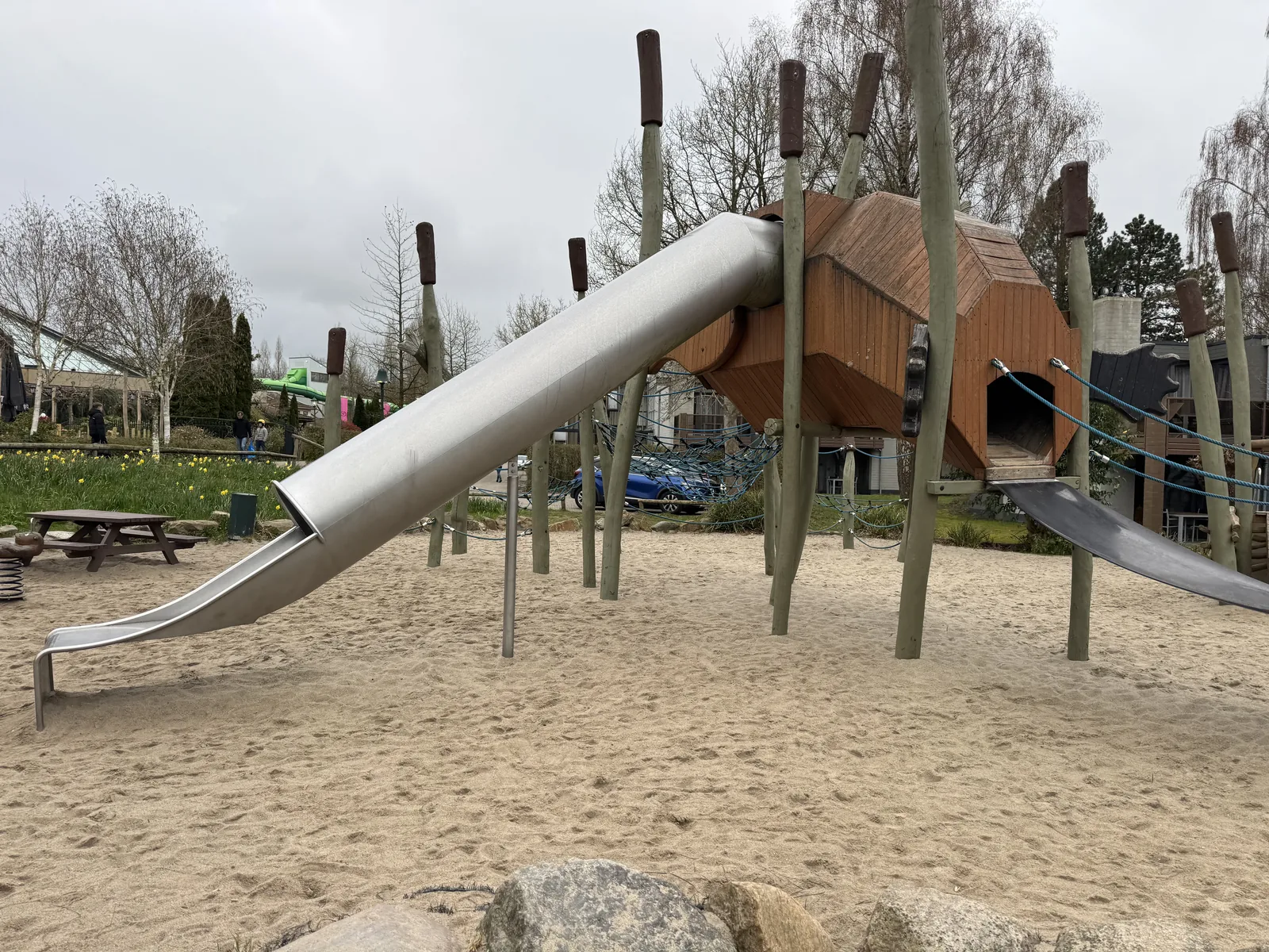 Spielplatz mit langer Edelstahlrutsche, hölzernem Kletterturm auf Pfählen und großer Sandfläche