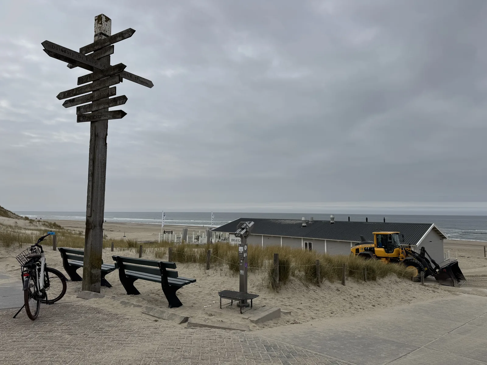 Hölzerner Wegweiserpfahl am Strandaufgang mit Bank und Strandpavillon im Hintergrund